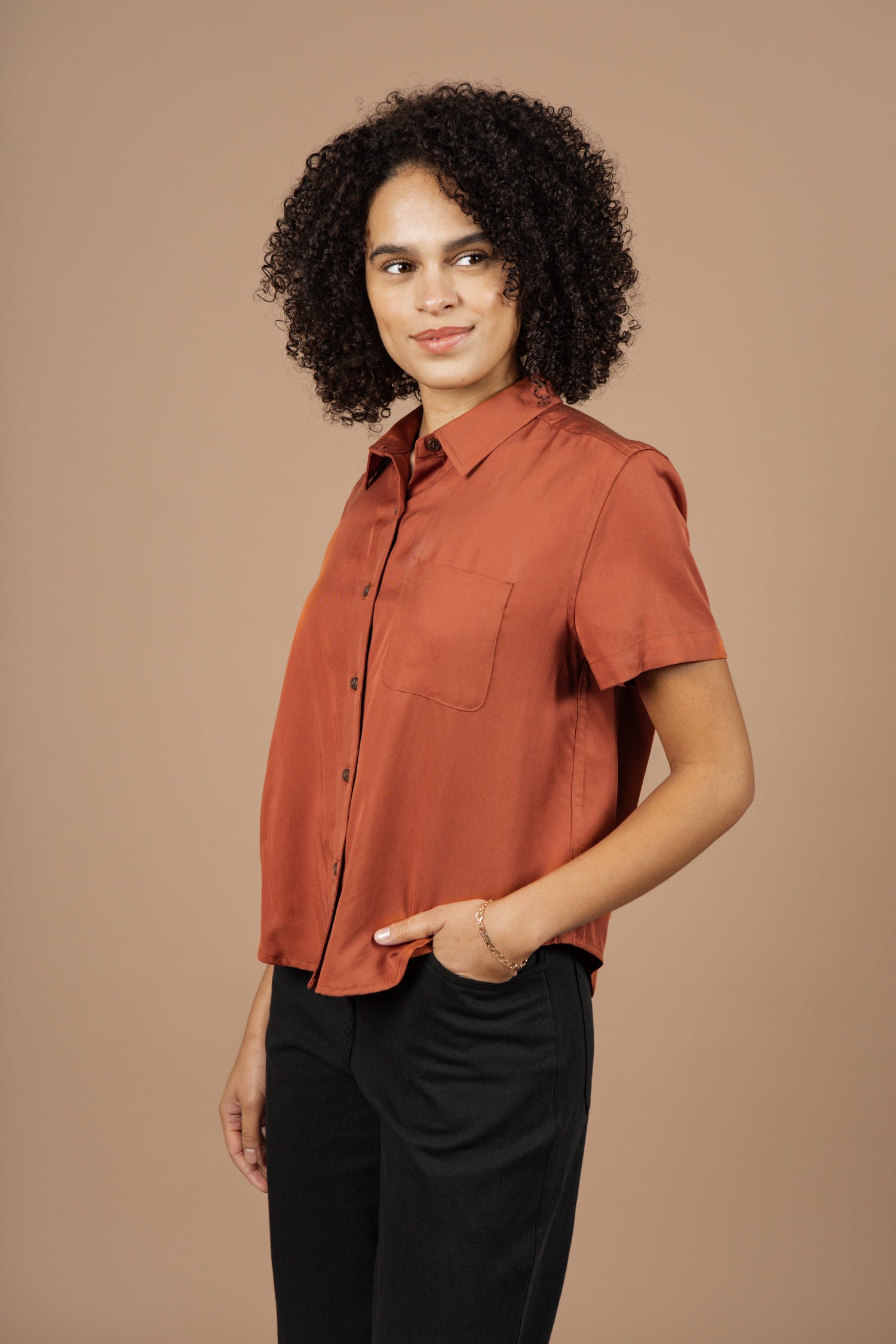 A woman with curly hair models the Bridge & Burn Greer Cropped Shirt in Copper—a boxy-fit Tencel top—paired with black pants. She stands against a beige background, glancing sideways with one hand in her pocket.
