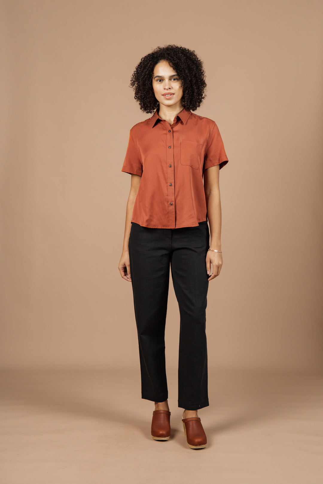 A woman with curly hair stands against a beige background, wearing the Bridge & Burn Greer Cropped Shirt in Copper, black pants, and brown shoes. She faces forward with a neutral expression.