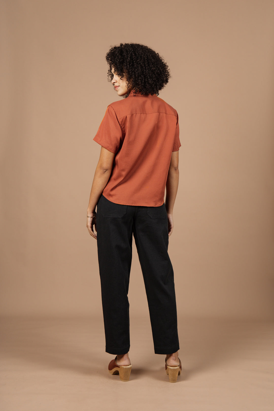 A person with curly hair stands facing away, wearing the Greer Cropped Shirt in copper by Bridge & Burn, paired with black pants and tan heeled sandals, against a plain beige background.