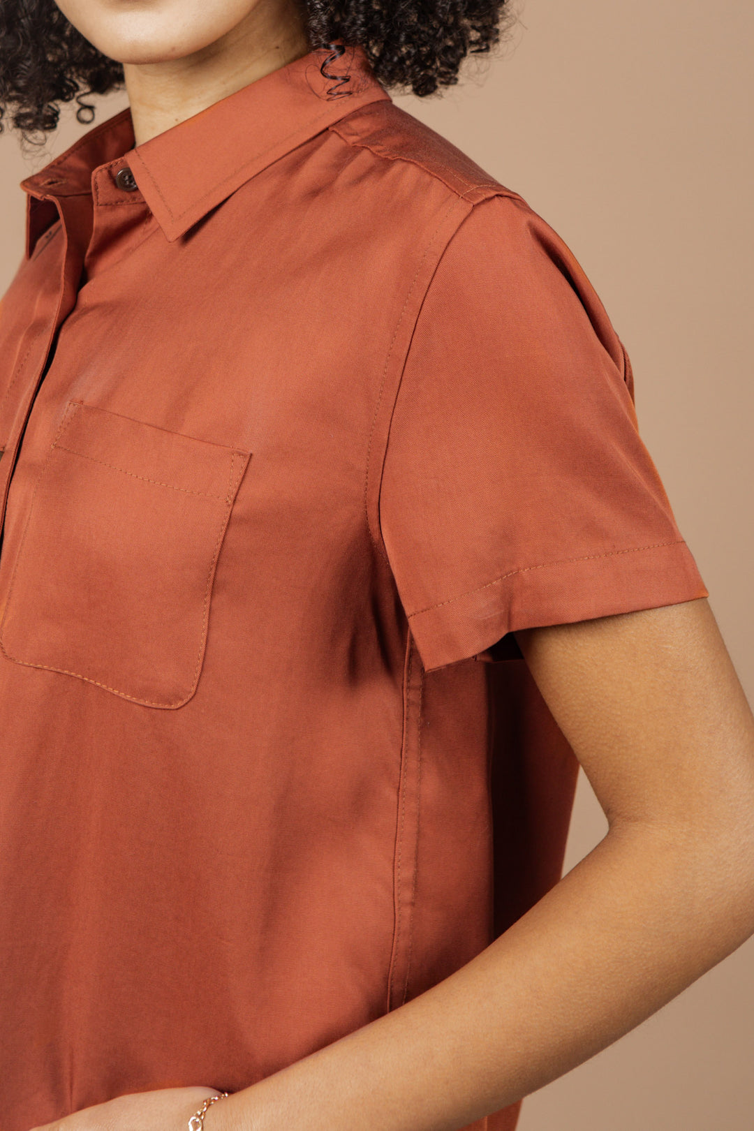 A person wearing the Bridge & Burn Greer Cropped Shirt in Copper stands against a beige background. The shirt has a boxy fit, only the upper body is visible, one arm is slightly bent, and the face is partially out of frame.