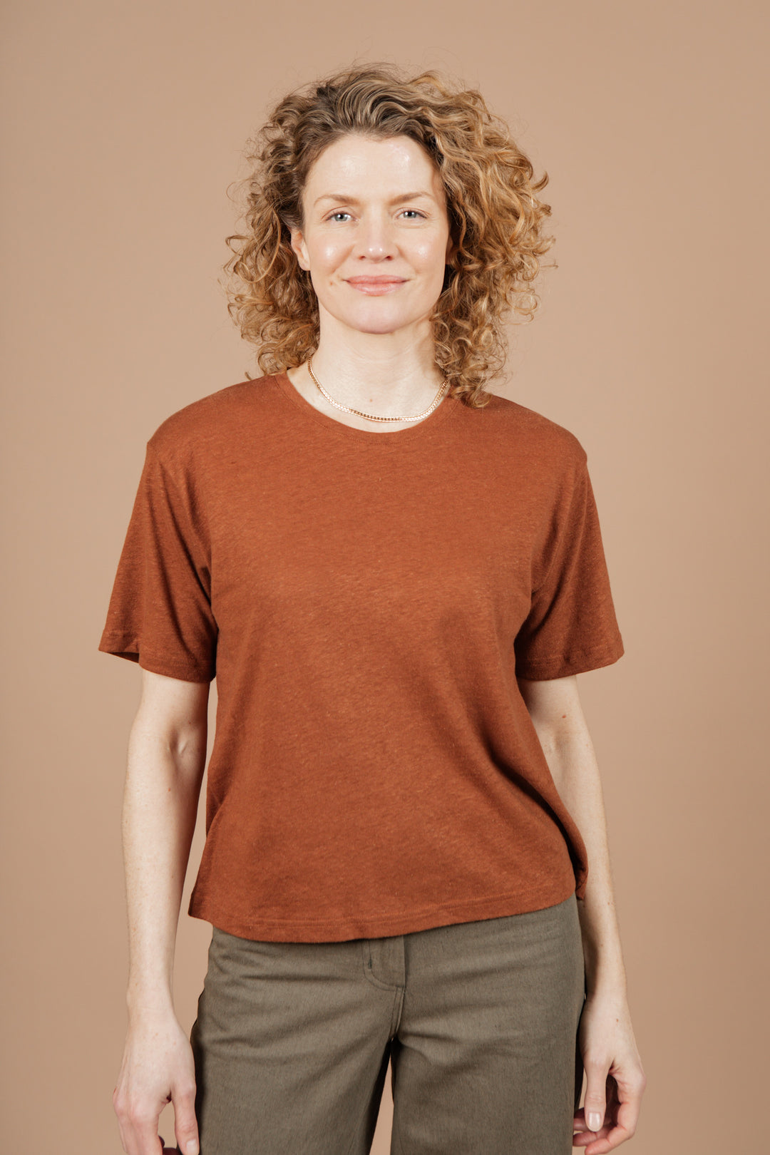 A person with curly, shoulder-length hair smiles while standing against a neutral beige background. They're wearing Bridge & Burn's Organic Hemp Boxy Tee in Burnt Umber and olive green pants.