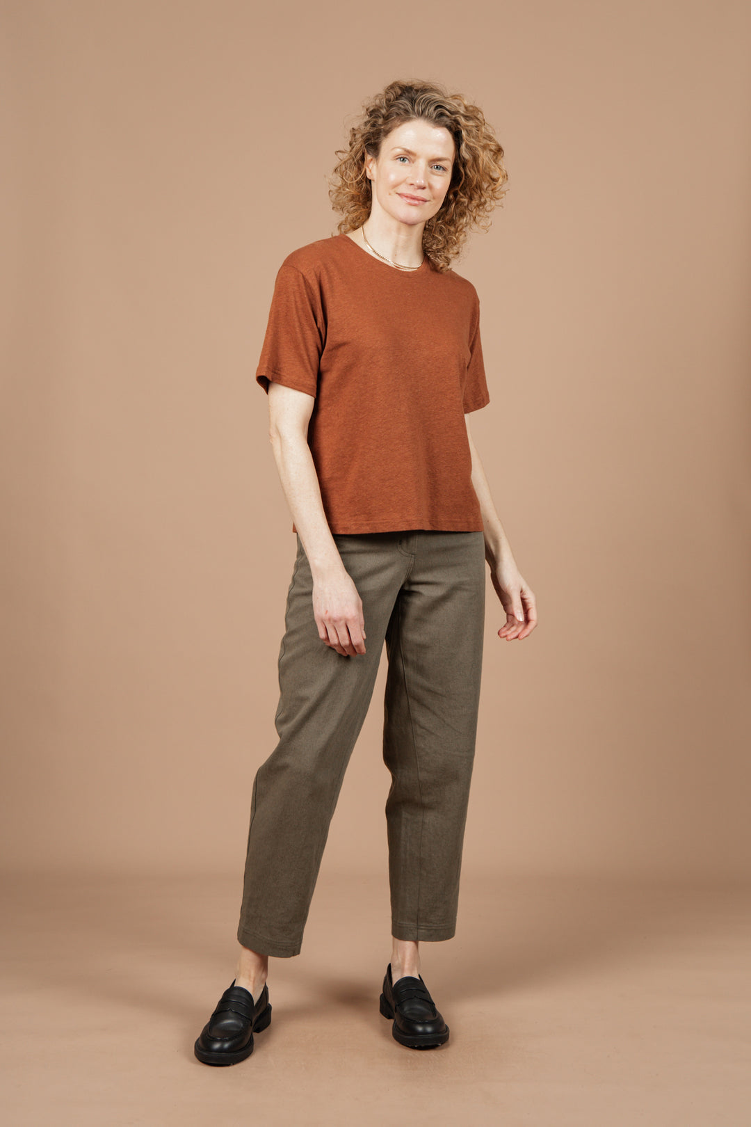 A person with curly hair stands against a beige background, wearing Bridge & Burn's Organic Hemp Boxy Tee in Burnt Umber, paired with olive green pants and black loafers, looking at the camera with a slight smile.