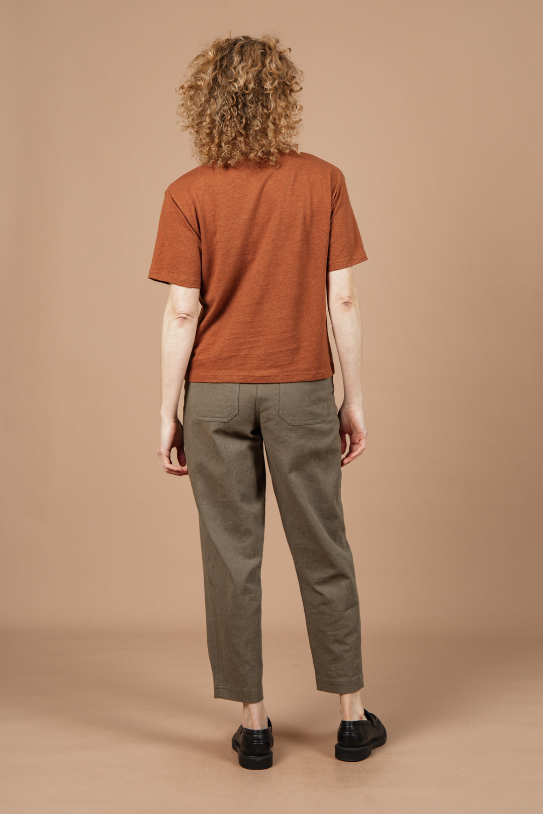 A person with curly hair faces away, wearing Bridge & Burn's Organic Hemp Boxy Tee in Burnt Umber, olive green pants, and black shoes against a plain beige background.
