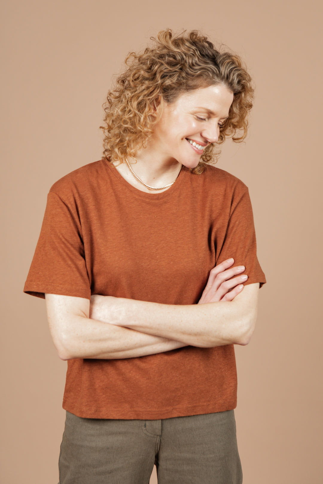 A person with curly, light brown hair wears the Bridge & Burn Organic Hemp Boxy Tee in Burnt Umber, standing against a beige background, smiling and looking down with arms crossed.