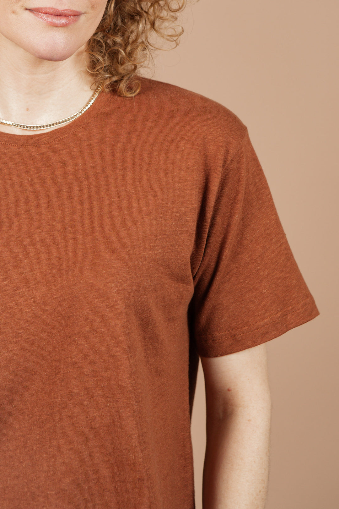 Close-up of a person in the Bridge & Burn Organic Hemp Boxy Tee in Burnt Umber, layered with a thin necklace and showing curly hair. Only the upper torso and part of the face appear against a plain, light brown background.