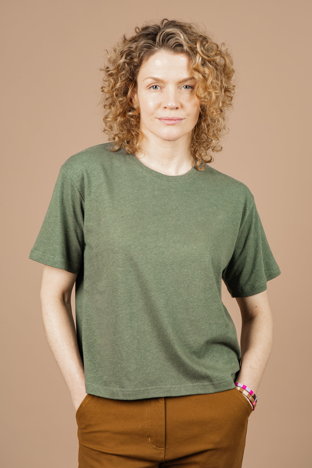 A person with curly, shoulder-length blonde hair stands against a tan background, wearing the Bridge & Burn Organic Hemp Boxy Tee in Cypress and brown pants. Their hands are in their pockets as they look neutrally at the camera.