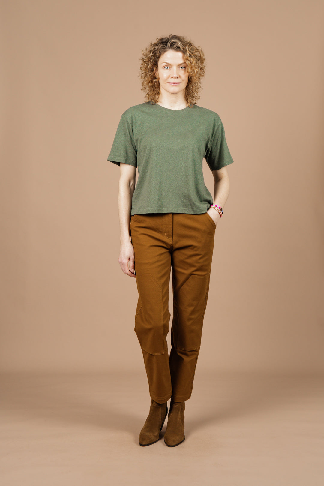 A person with curly hair faces forward against a plain beige background, wearing the Bridge & Burn Organic Hemp Boxy Tee in Cypress, paired with brown pants and brown boots.