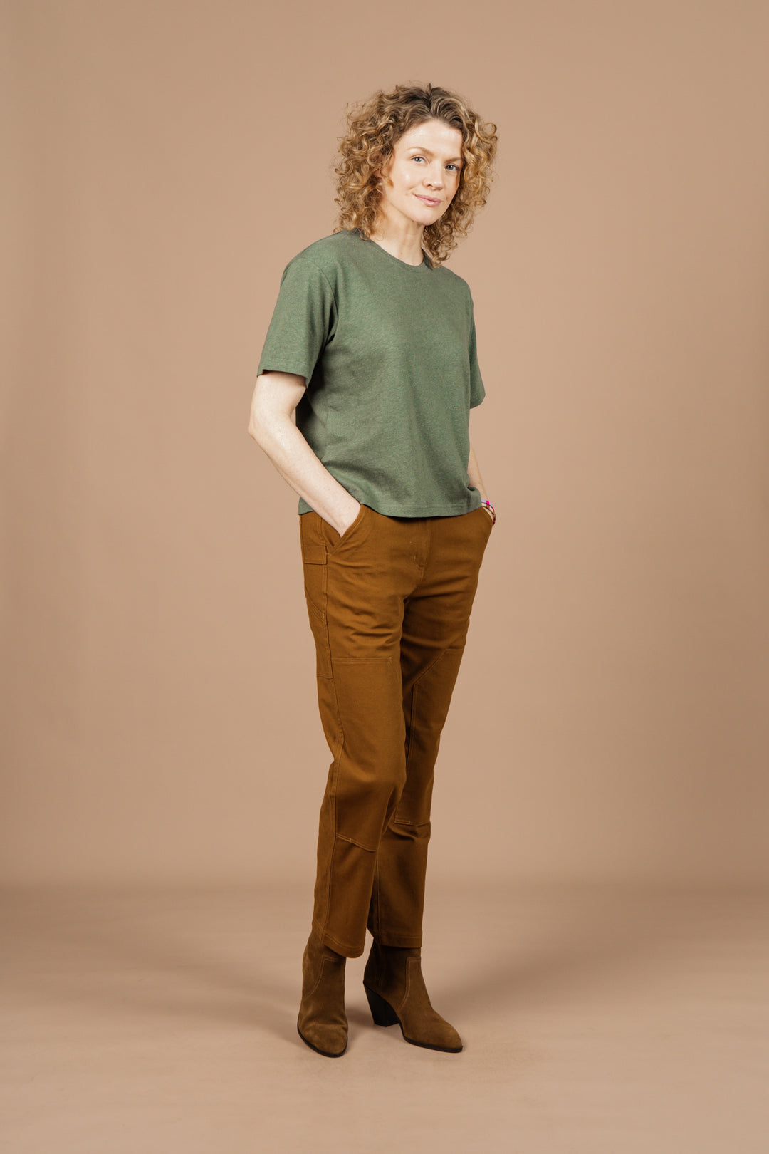A person with curly hair stands against a beige background, wearing Bridge & Burn’s Organic Hemp Boxy Tee in Cypress, brown pants, and brown ankle boots, hands in their pockets and looking relaxed.