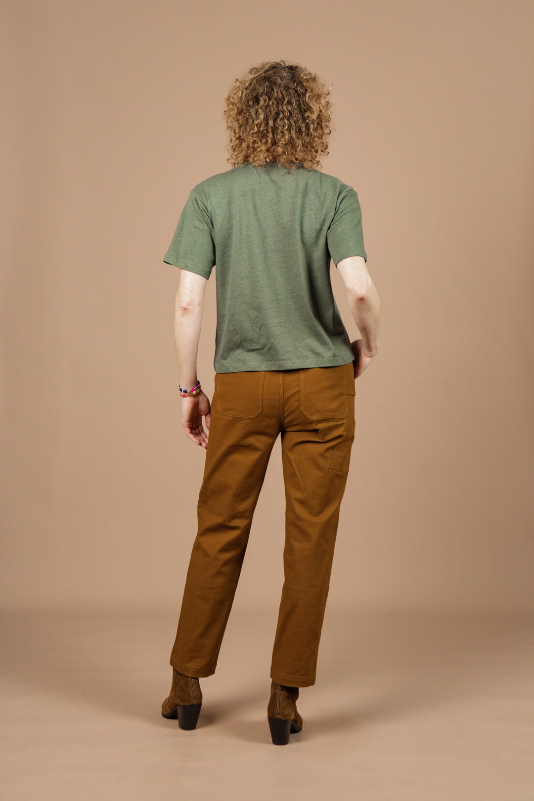 A person with curly hair, seen from behind, wears the Bridge & Burn Organic Hemp Boxy Tee in Cypress, paired with brown pants and brown shoes, standing against a plain beige background.
