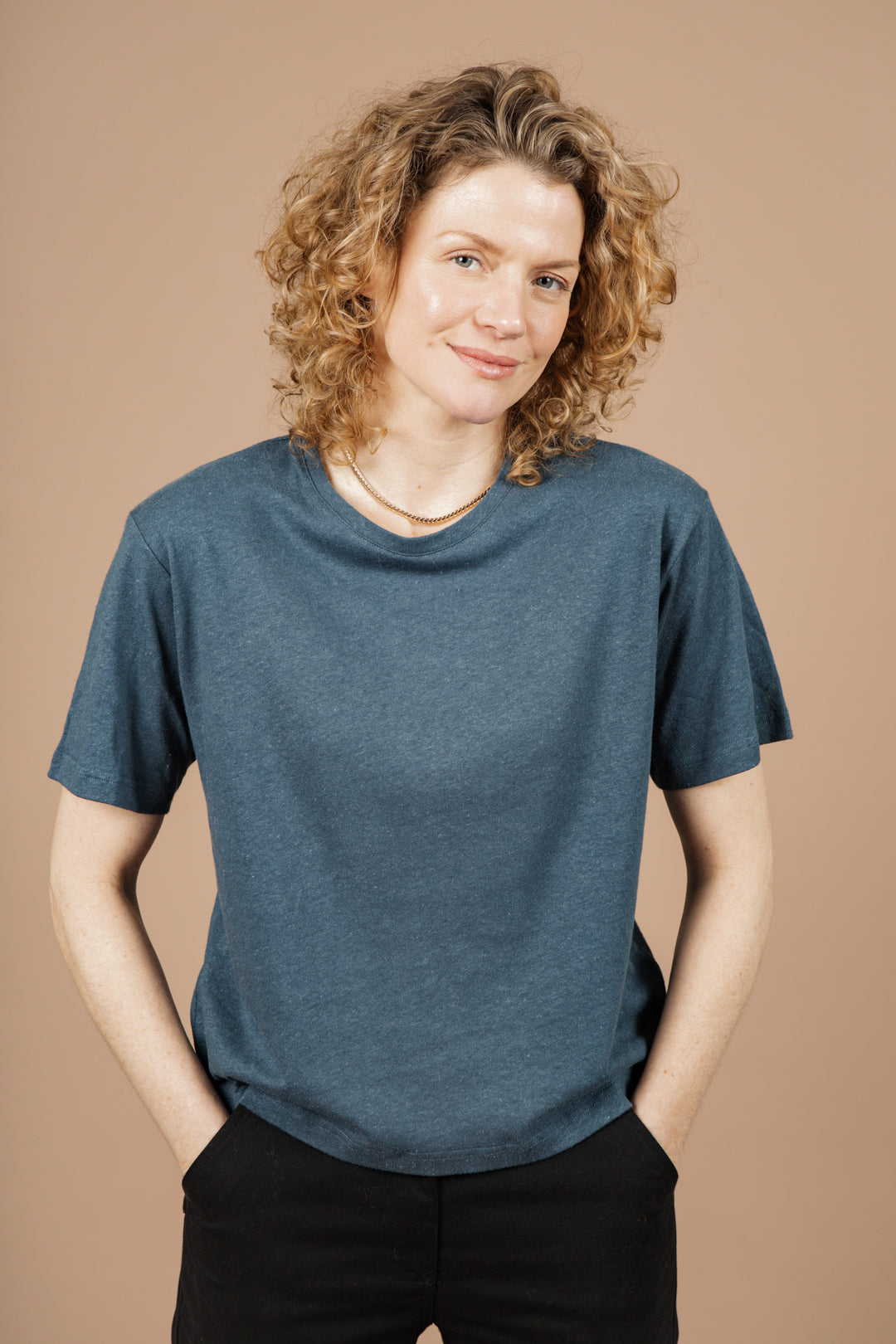 A person with curly blonde hair wears the Bridge & Burn Organic Hemp Boxy Tee in Storm Blue and black pants, standing against a beige background, smiling gently with hands in pockets.