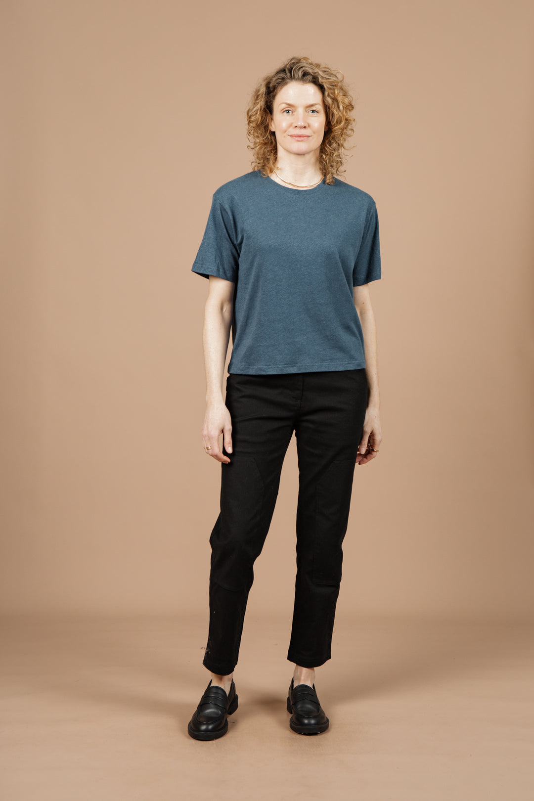 A person with curly blonde hair wears the Bridge & Burn Organic Hemp Boxy Tee in Storm Blue, paired with black pants and black shoes, standing facing forward against a plain beige background.