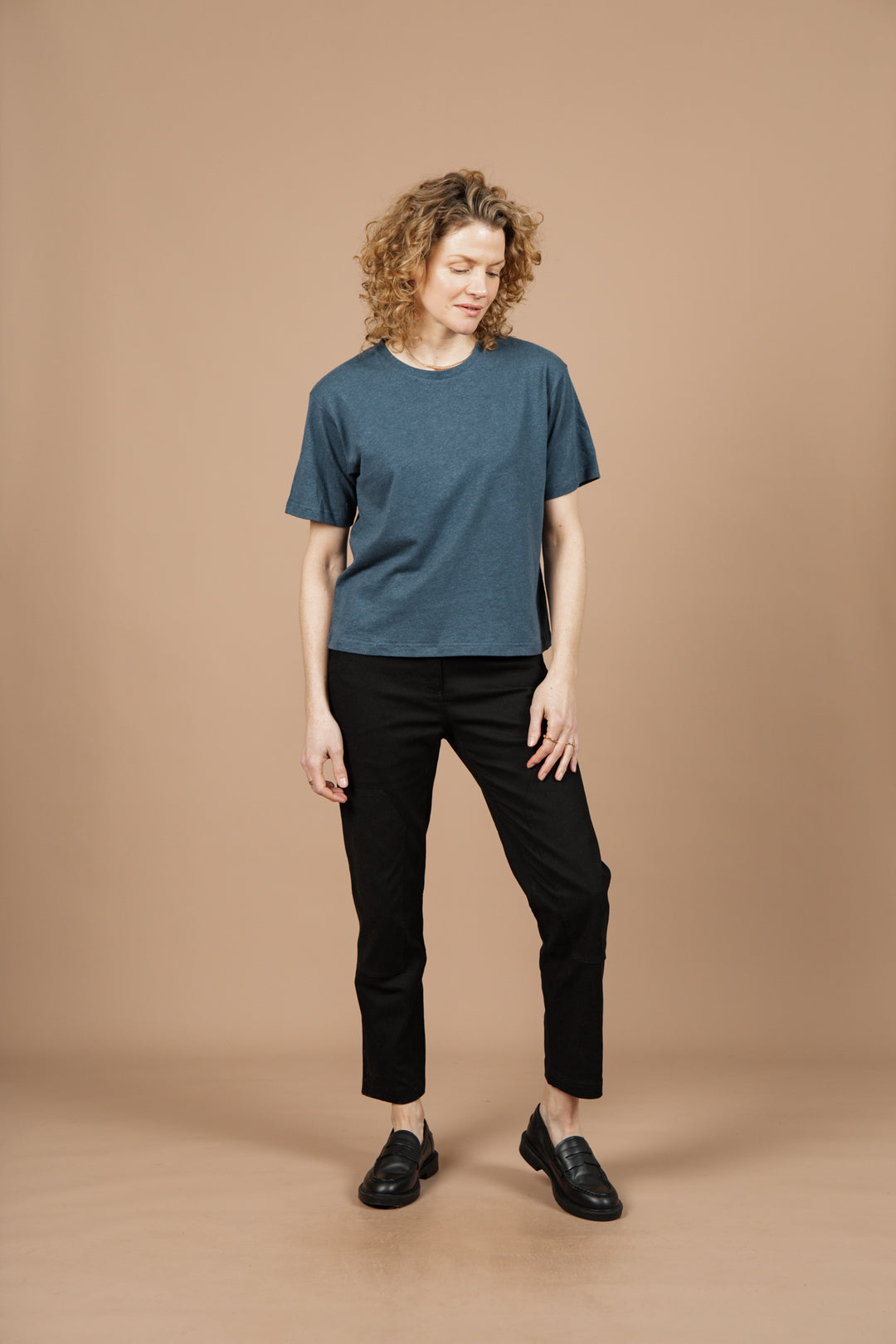 A person with curly blond hair wears the Bridge & Burn Organic Hemp Boxy Tee in Storm Blue, paired with black pants and shoes, standing against a beige background and looking down with a neutral expression.