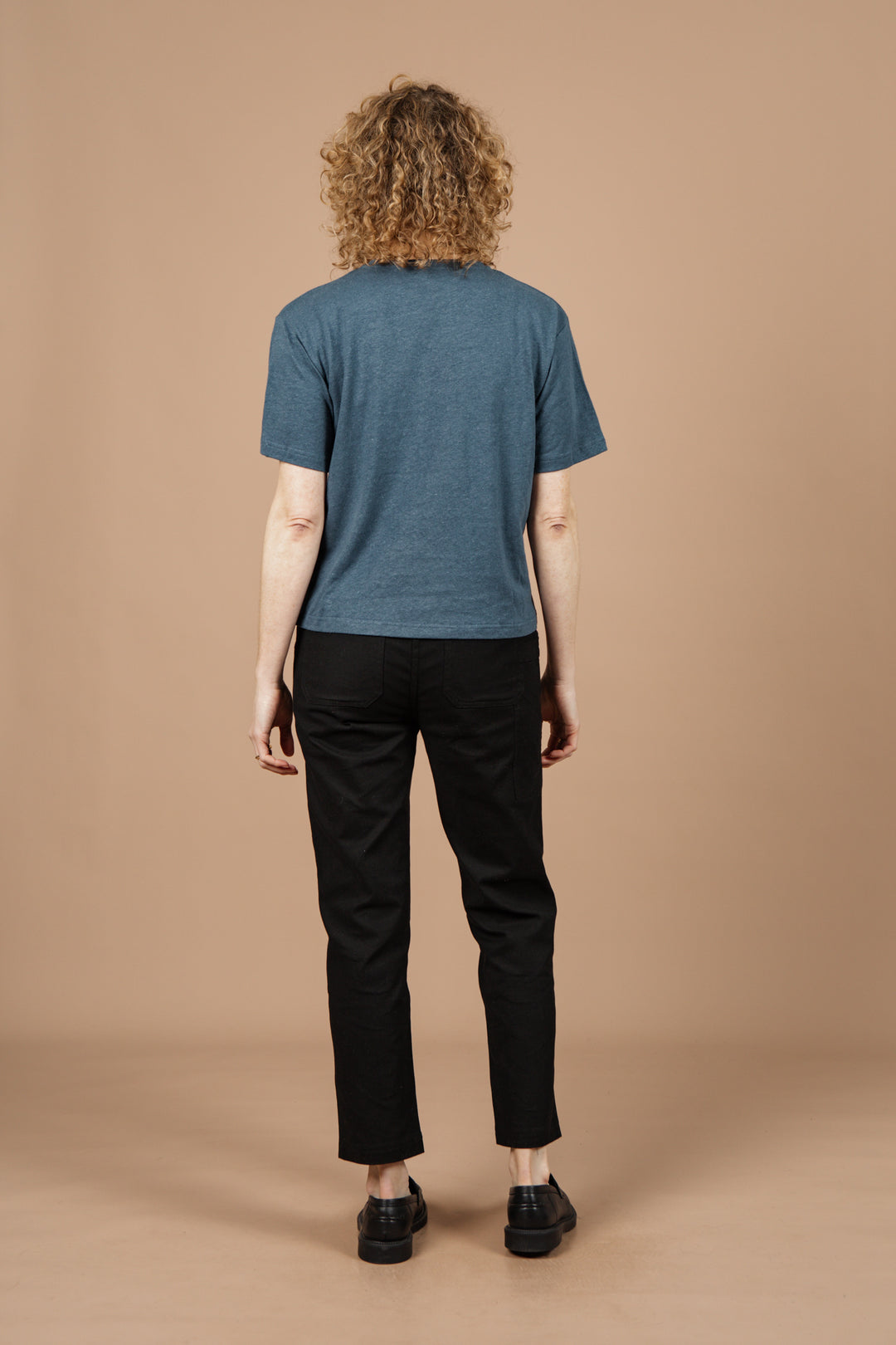 A person with curly hair faces away from the camera wearing the Bridge & Burn Organic Hemp Boxy Tee in Storm Blue, black pants, and black shoes, set against a plain beige background.