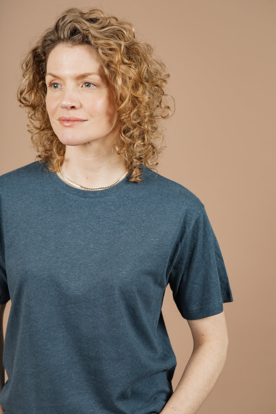 A woman with curly blonde hair wears the Bridge & Burn Organic Hemp Boxy Tee in Storm Blue and a necklace, standing against a plain beige background, gazing slightly to the side with a neutral expression.