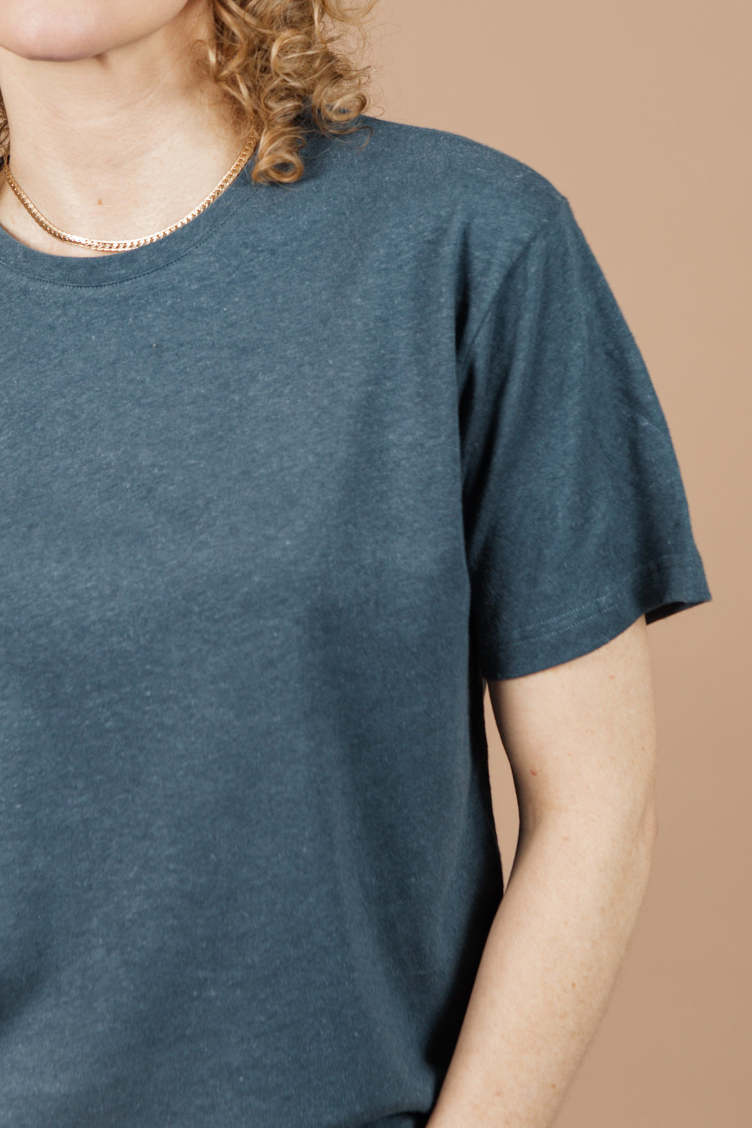 A person with curly hair, face not shown, is partially visible against a beige background wearing the Bridge & Burn Organic Hemp Boxy Tee in Storm Blue and a gold chain necklace.