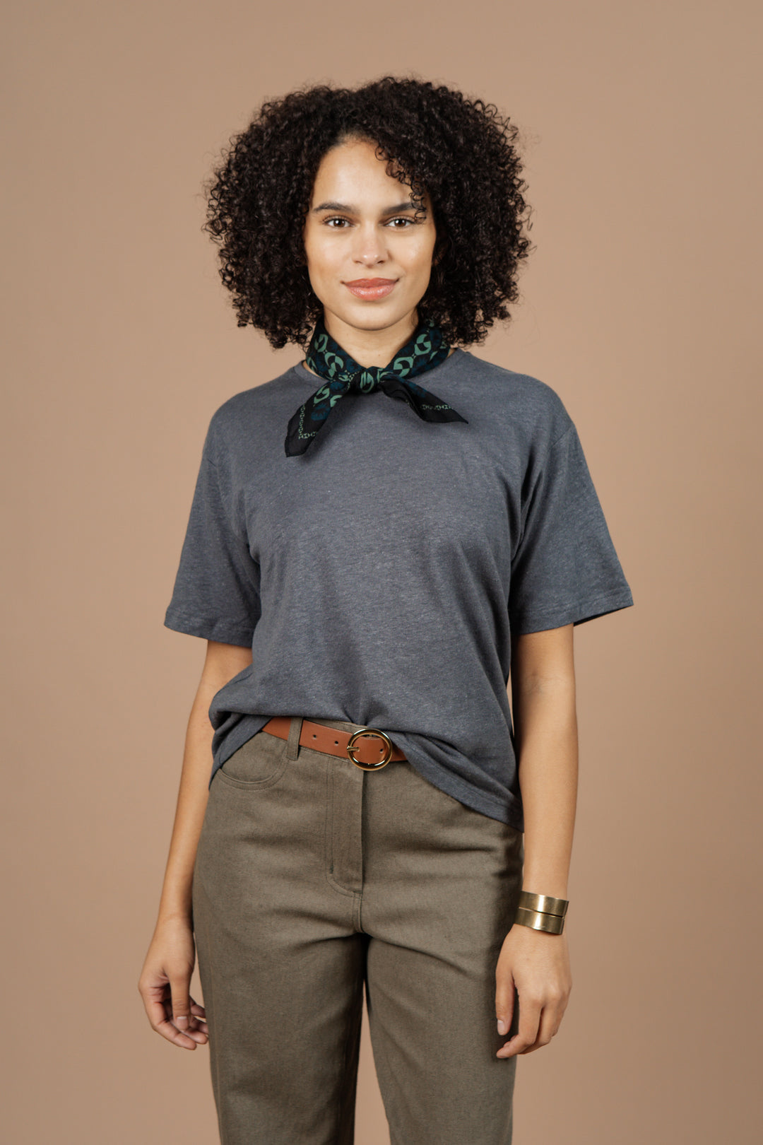 A person with curly hair wears Bridge & Burn's Organic Hemp Boxy Tee in Slate Grey, styled with olive pants, a brown belt, green patterned scarf, and gold bracelet, standing against a plain beige background.