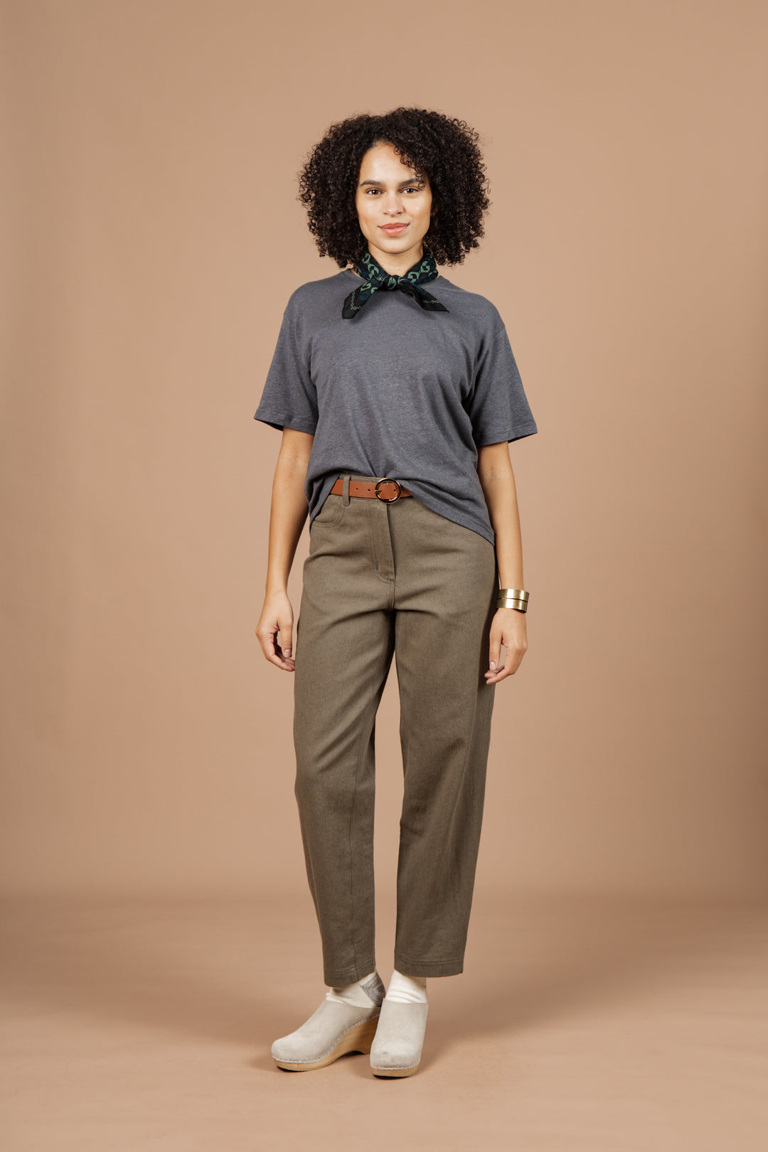 A woman with curly hair stands against a beige background, wearing the Bridge & Burn Organic Hemp Boxy Tee in Slate Grey, olive green pants with a brown belt, white shoes, a green patterned neck scarf, and a gold bracelet.