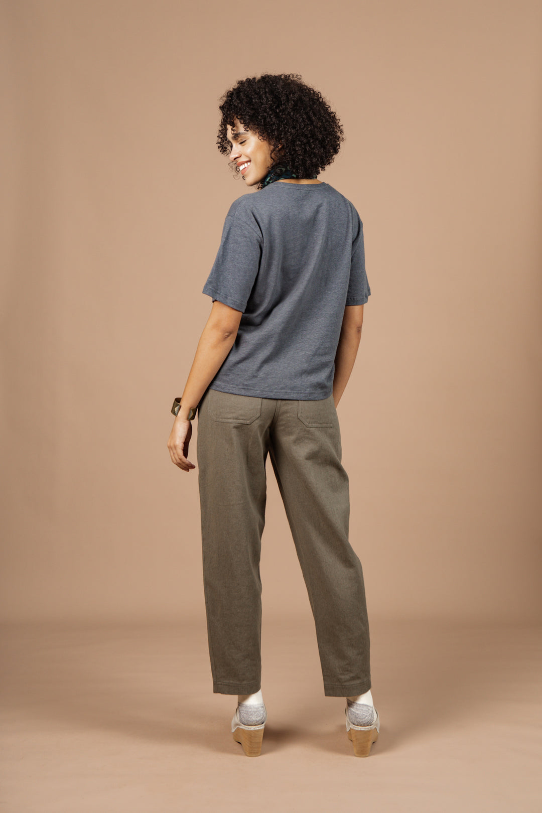 A woman with curly hair smiles over her shoulder, wearing the Bridge & Burn Organic Hemp Boxy Tee in Slate Grey, olive green pants, and light shoes against a solid beige background.