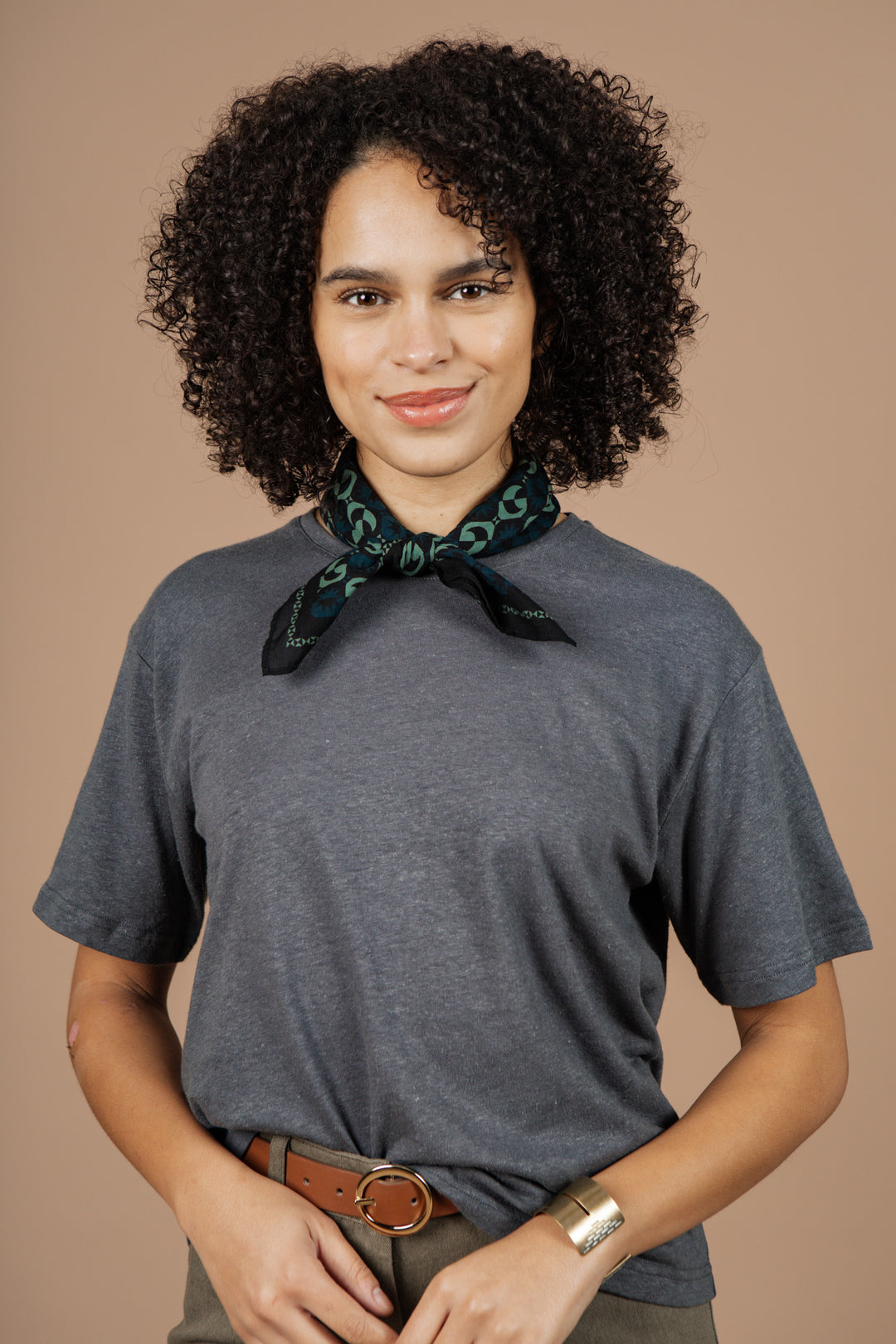 A woman with curly hair, wearing the Bridge & Burn Organic Hemp Boxy Tee in Slate Grey and a patterned scarf, brown belted pants, and a gold bracelet, stands against a beige background and smiles at the camera.