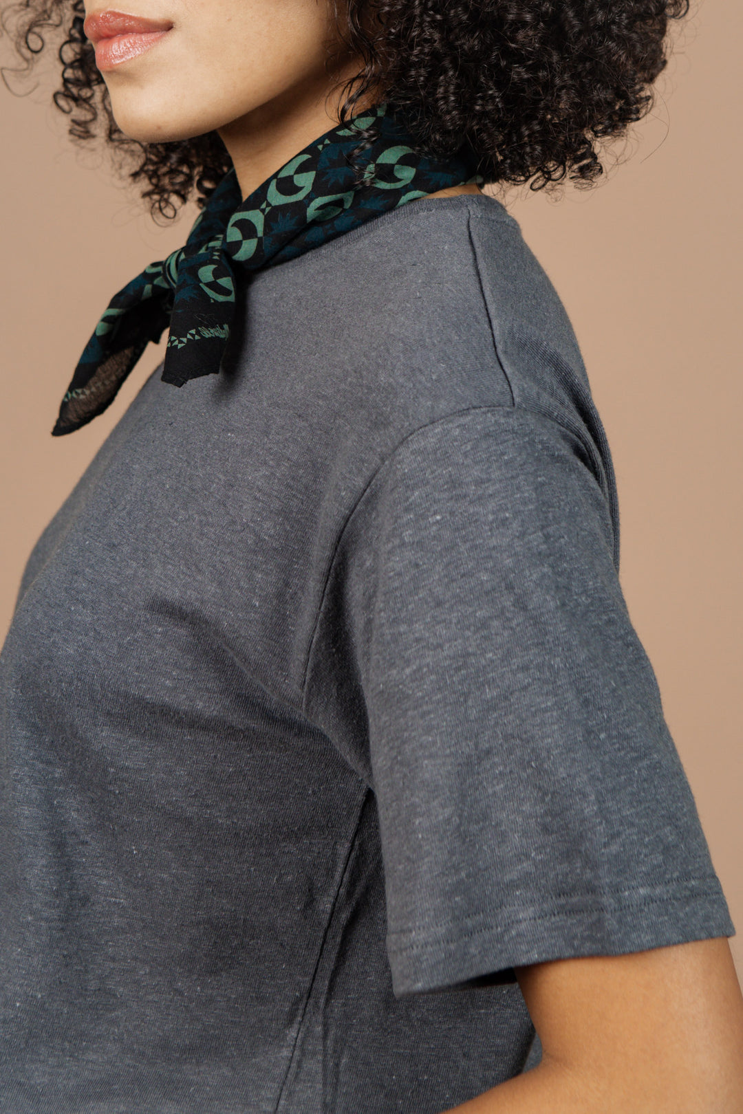 A person with curly hair wears the Bridge & Burn Organic Hemp Boxy Tee in Slate Grey and a black and green patterned scarf tied at their neck, shown from the side against a plain beige background.