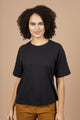 A person with curly hair wears the Bridge & Burn Organic Hemp Boxy Tee in Washed Black and brown pants, standing against a plain beige background and smiling softly at the camera.