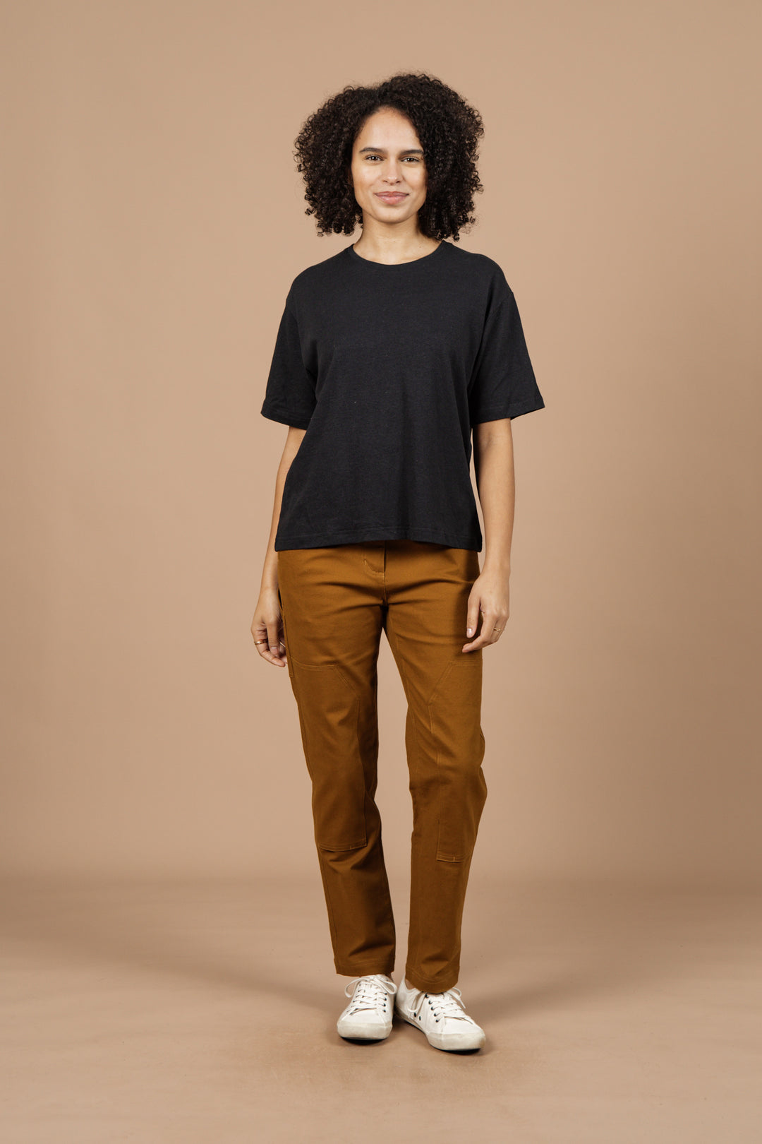 A person with curly hair stands against a beige background, facing forward with a slight smile, wearing the Bridge & Burn Organic Hemp Boxy Tee in Washed Black, paired with brown pants and white sneakers.