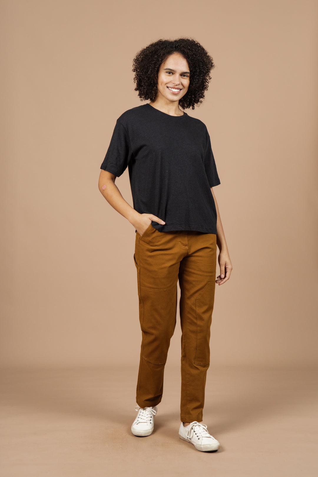 A person with curly hair is smiling, wearing the Bridge & Burn Organic Hemp Boxy Tee in Washed Black, brown pants, and white sneakers with one hand in their pocket against a beige background.