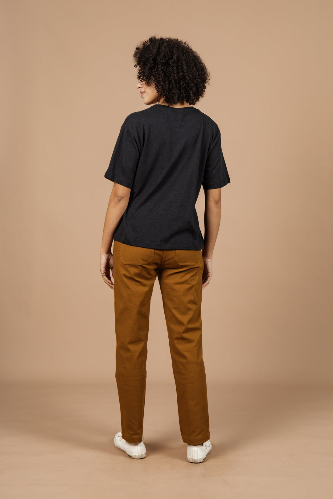 A person with curly hair stands facing away, wearing the Bridge & Burn Organic Hemp Boxy Tee in Washed Black, brown pants, and white sneakers against a plain beige background.