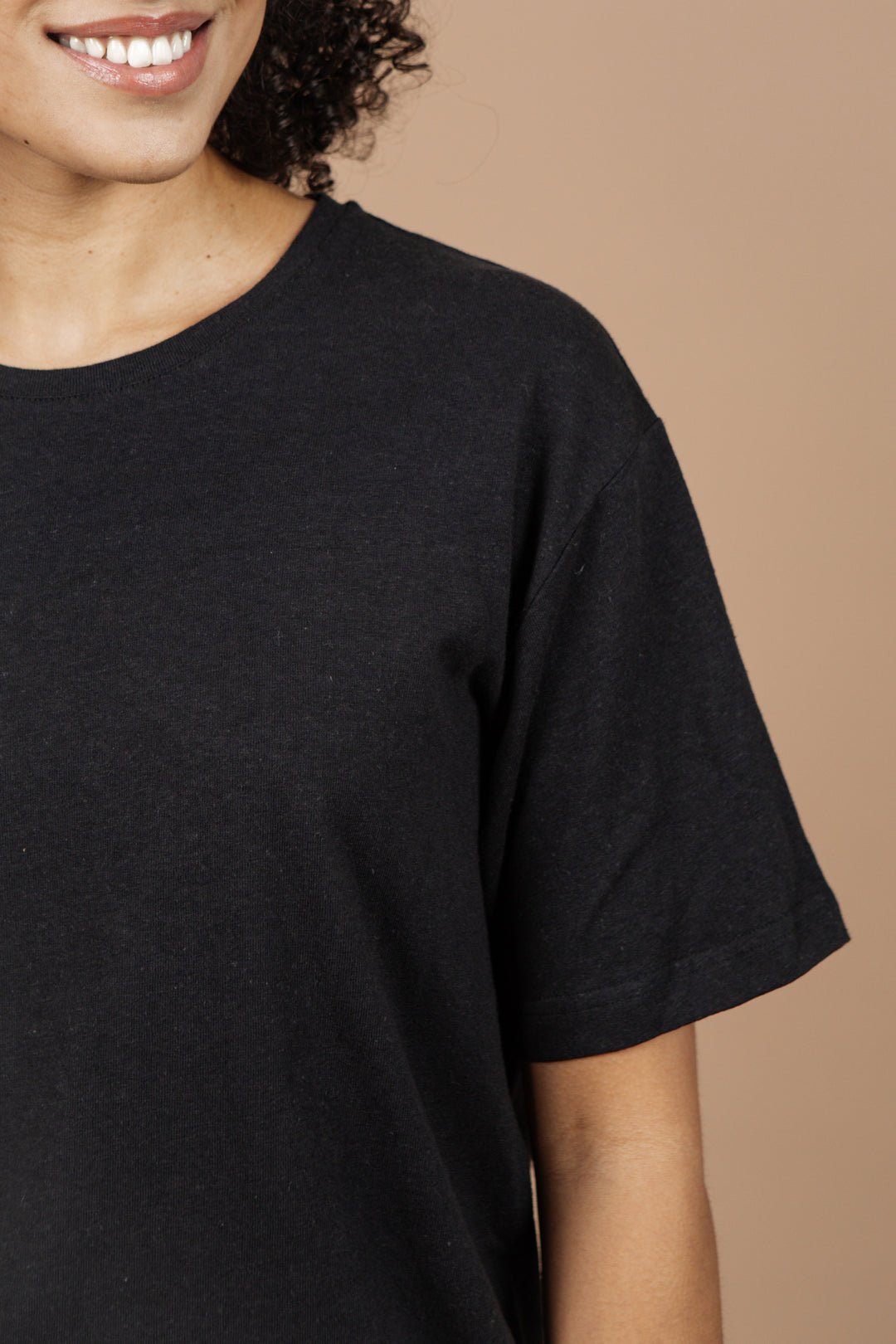 A woman with curly hair smiles, wearing the Bridge & Burn Organic Hemp Boxy Tee in Washed Black. Only her lower face, neck, shoulder, and upper chest are visible against a beige background.