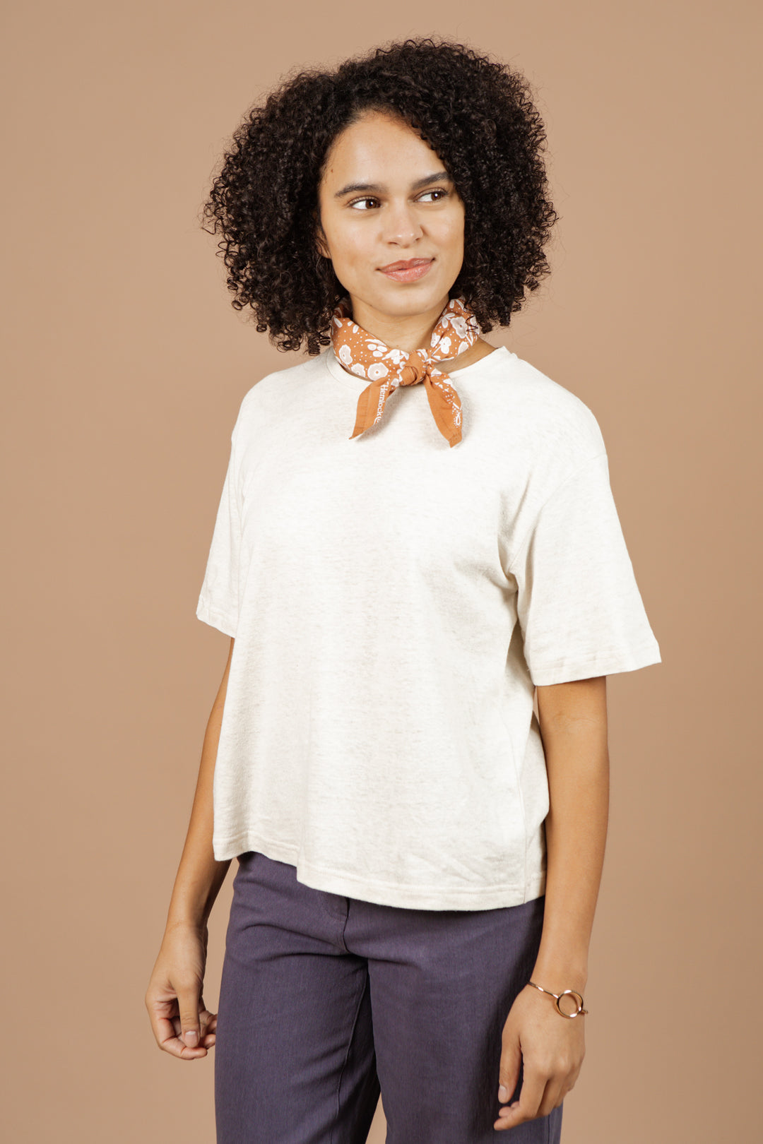 A woman with curly hair wears the Bridge & Burn Organic Hemp Boxy Tee in Ivory, dark pants, and an orange patterned scarf. She stands against a beige background, looking slightly to the side and smiling.