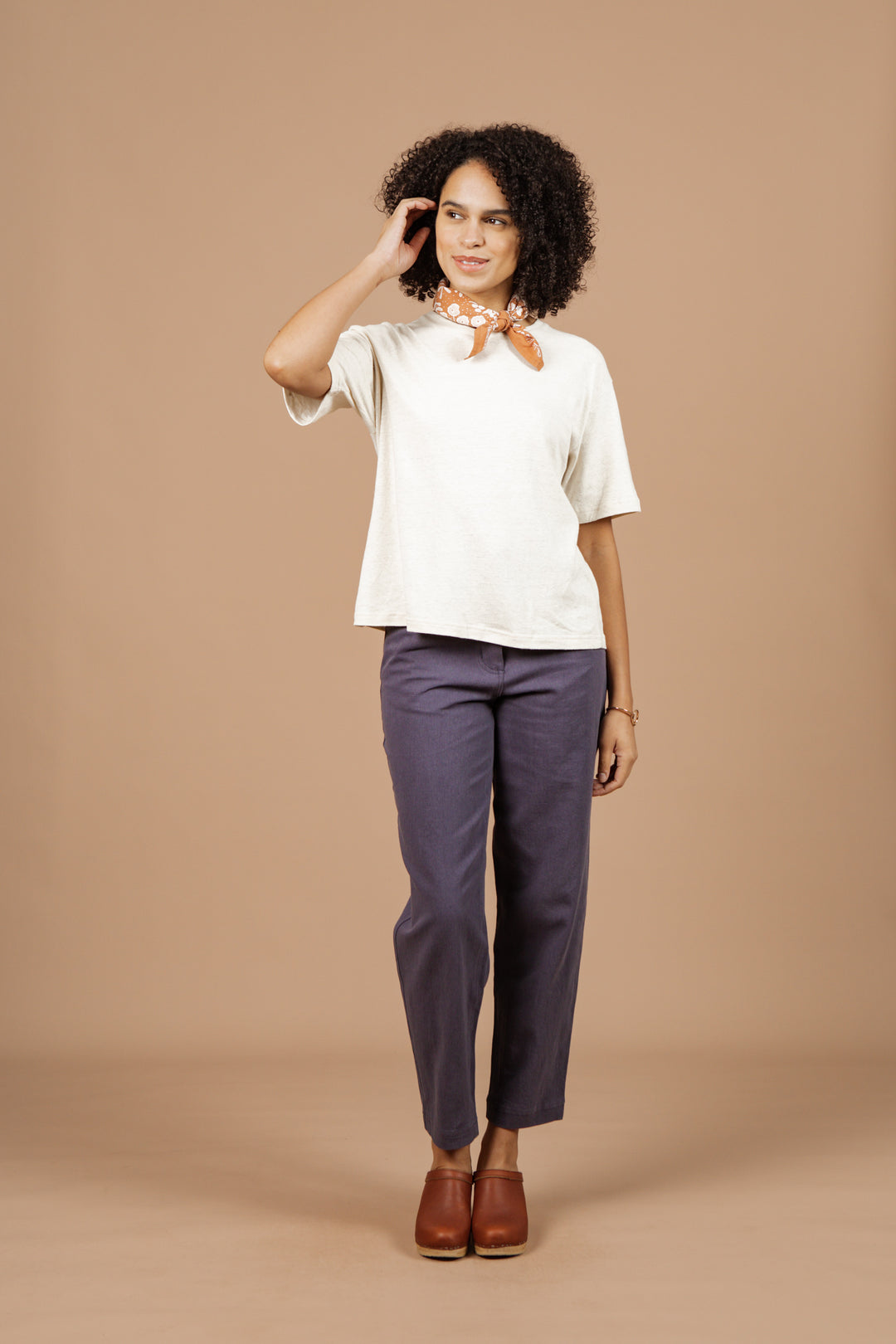 A woman with curly hair stands against a beige background, smiling as she gently touches her hair. She wears the Bridge & Burn Organic Hemp Boxy Tee in Ivory, paired with blue pants and brown accessories for an effortlessly chic look.