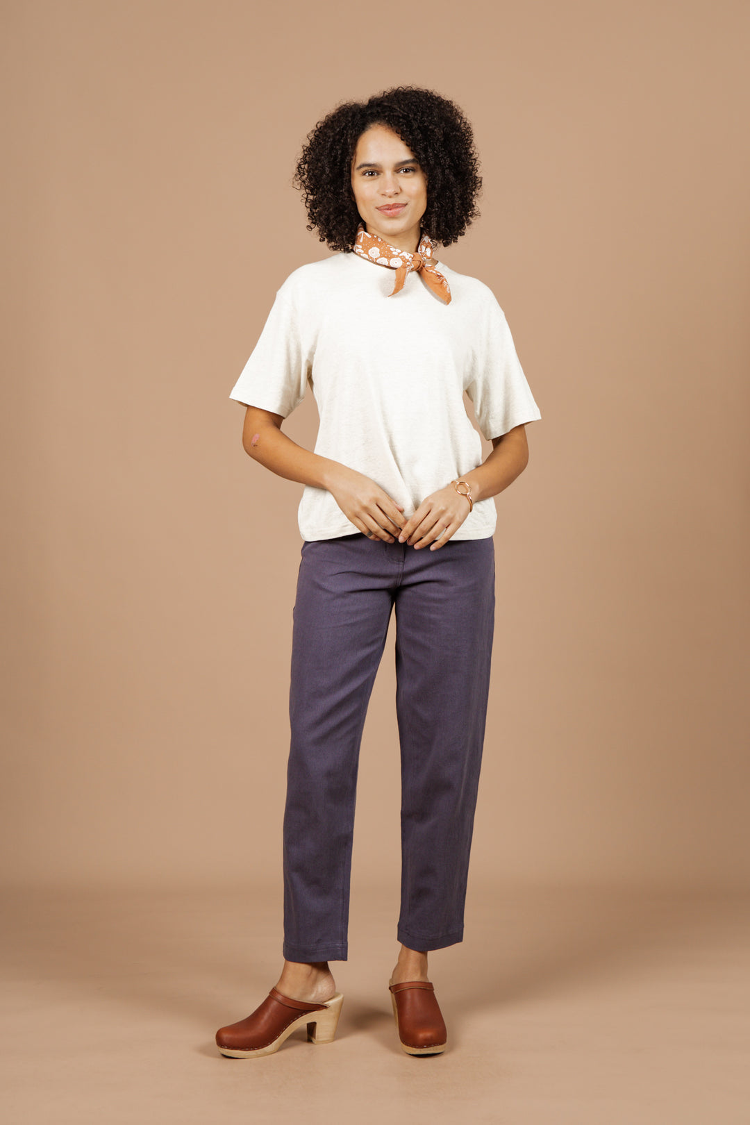 A person with curly hair faces forward in an ivory Organic Hemp Boxy Tee by Bridge & Burn, blue pants, brown clogs, and a light scarf, set against a beige background.