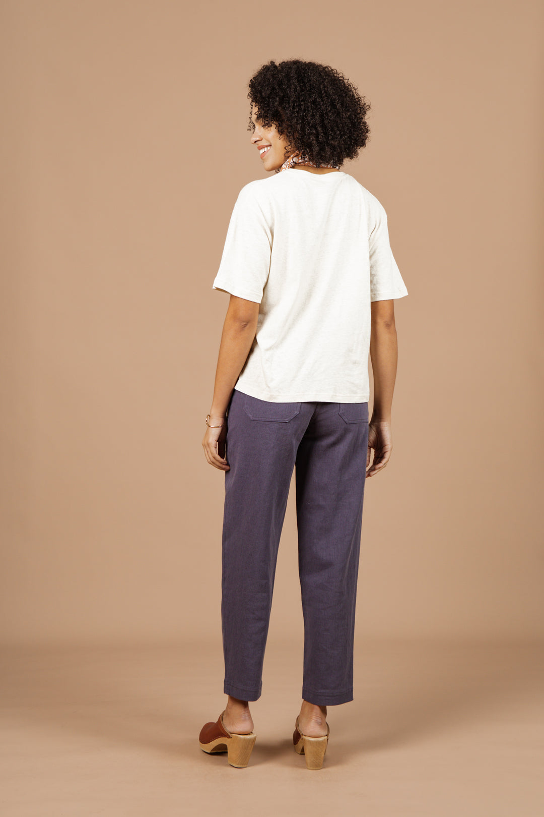 A woman smiles over her shoulder, wearing the Bridge & Burn Organic Hemp Boxy Tee in Ivory, paired with loose-fitting blue pants and tan heeled sandals. She stands against a plain, light brown background.