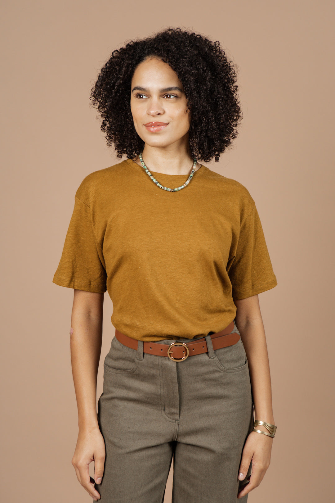 A woman with curly hair wears the Bridge & Burn Organic Hemp Boxy Tee in Ochre, grayish-brown pants, a brown belt, a beaded necklace, and a bracelet, standing against a plain beige background.