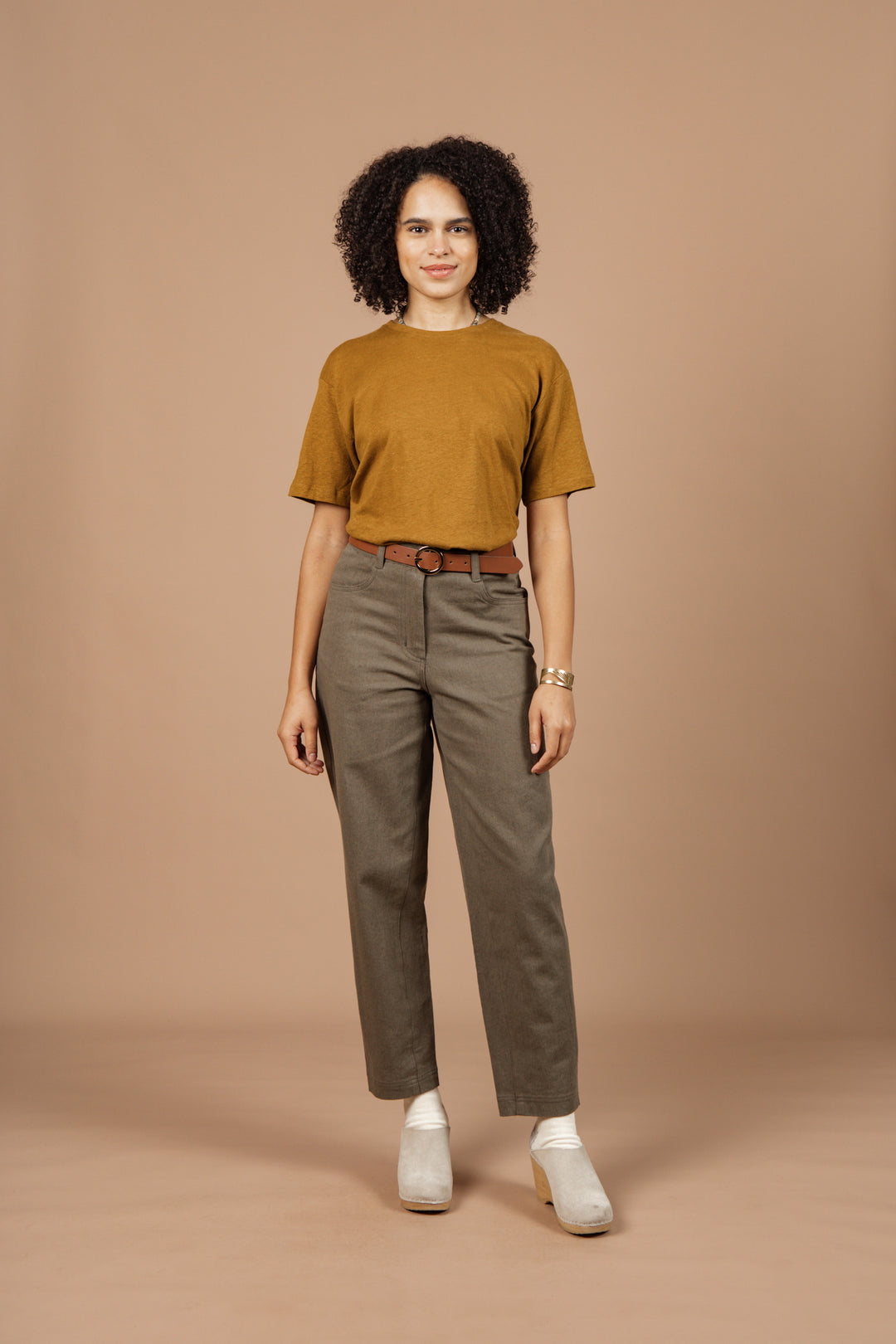 A woman with curly hair stands against a beige background, wearing the Bridge & Burn Organic Hemp Boxy Tee in ochre, high-waisted brown belted pants, white heeled shoes, and a watch. She smiles slightly and faces forward.