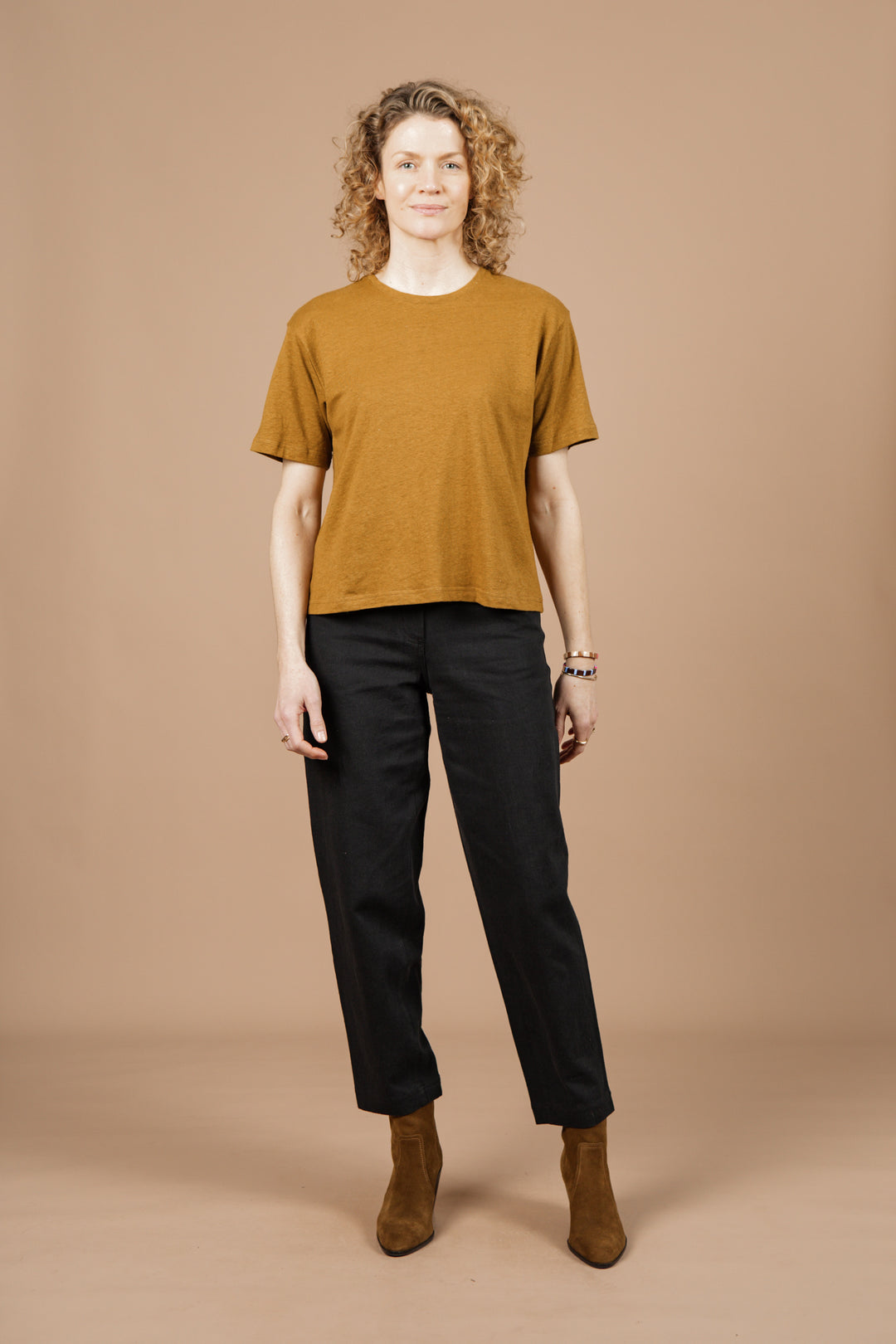 A person with curly hair faces forward against a beige background, wearing the Bridge & Burn Organic Hemp Boxy Tee in Ochre, paired with black pants and brown boots.