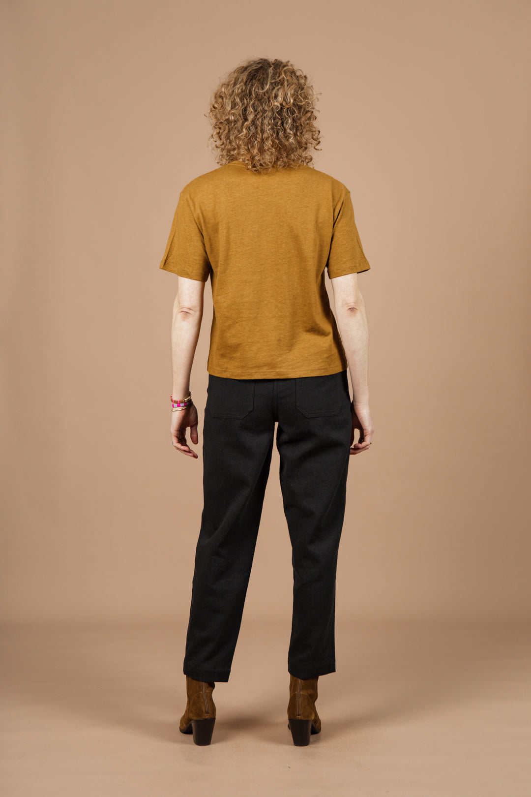 A person with curly hair faces away, wearing the Bridge & Burn Organic Hemp Boxy Tee in Ochre with black pants and brown heeled shoes, standing against a plain beige background.