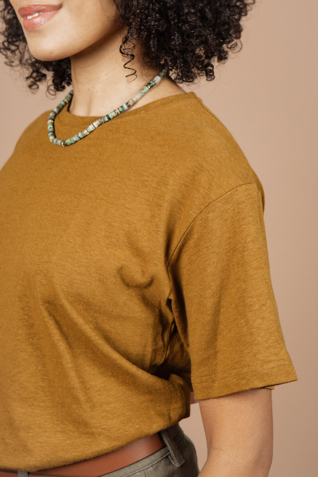 A woman with curly hair wears the Bridge & Burn Organic Hemp Boxy Tee in ochre and a green beaded necklace, shown from the shoulders up against a beige background.