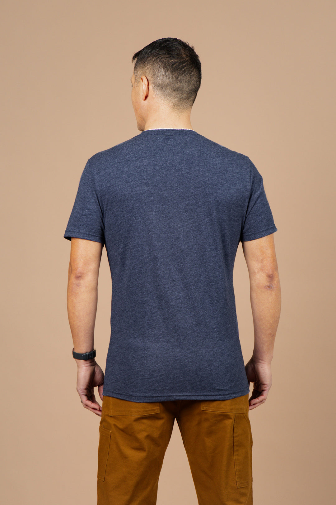 A man with short dark hair, wearing the Bridge & Burn Men's Alpenglow Tee in navy and brown pants, stands facing away from the camera against a plain beige background.