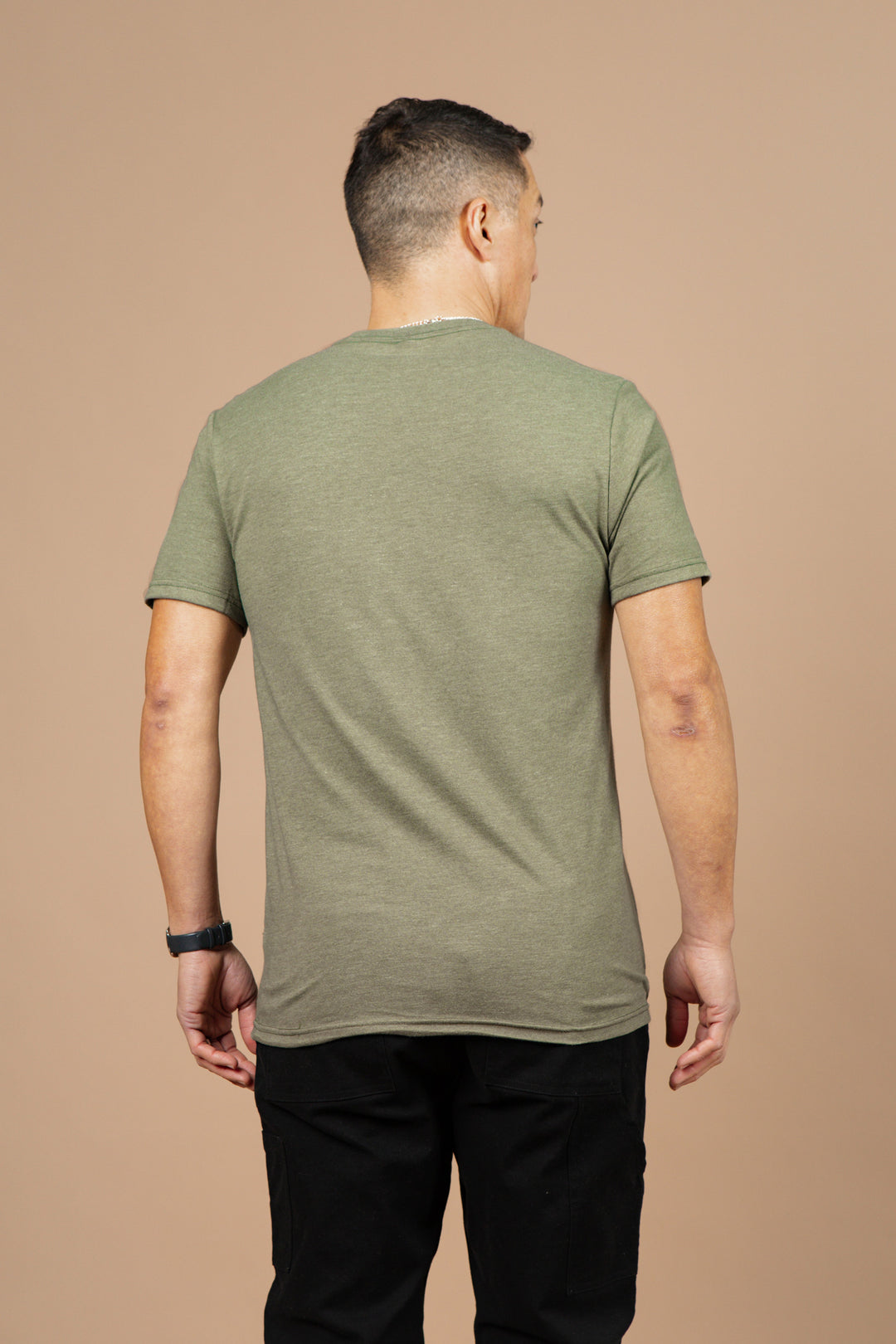 A man wearing the Bridge & Burn Men's Flown Tee in olive and black pants stands with his back to the camera against a plain beige background.