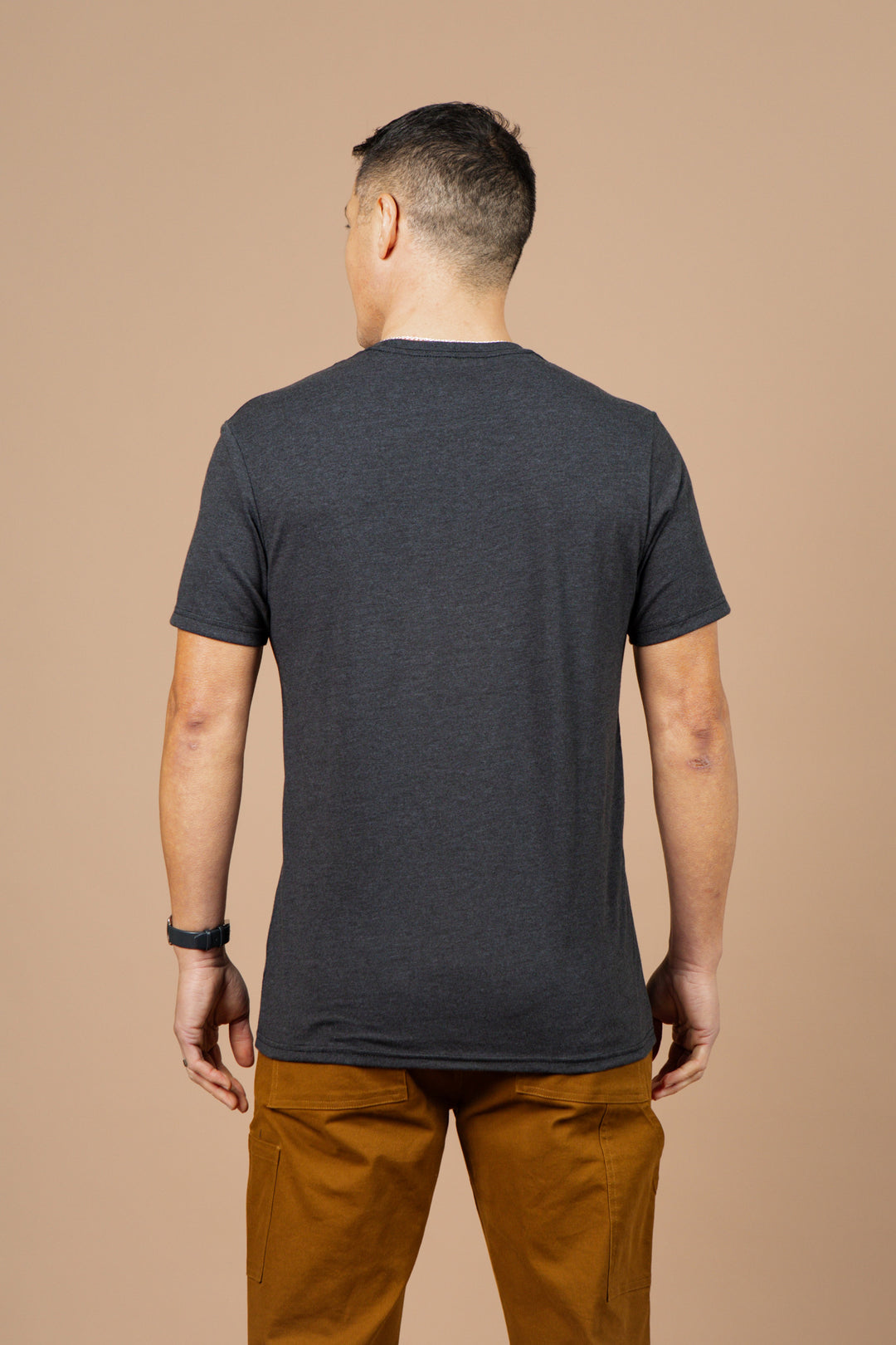 A man stands with his back to the camera, wearing the Bridge & Burn Men's Night Watch Tee in black and tan pants, set against a plain beige background.