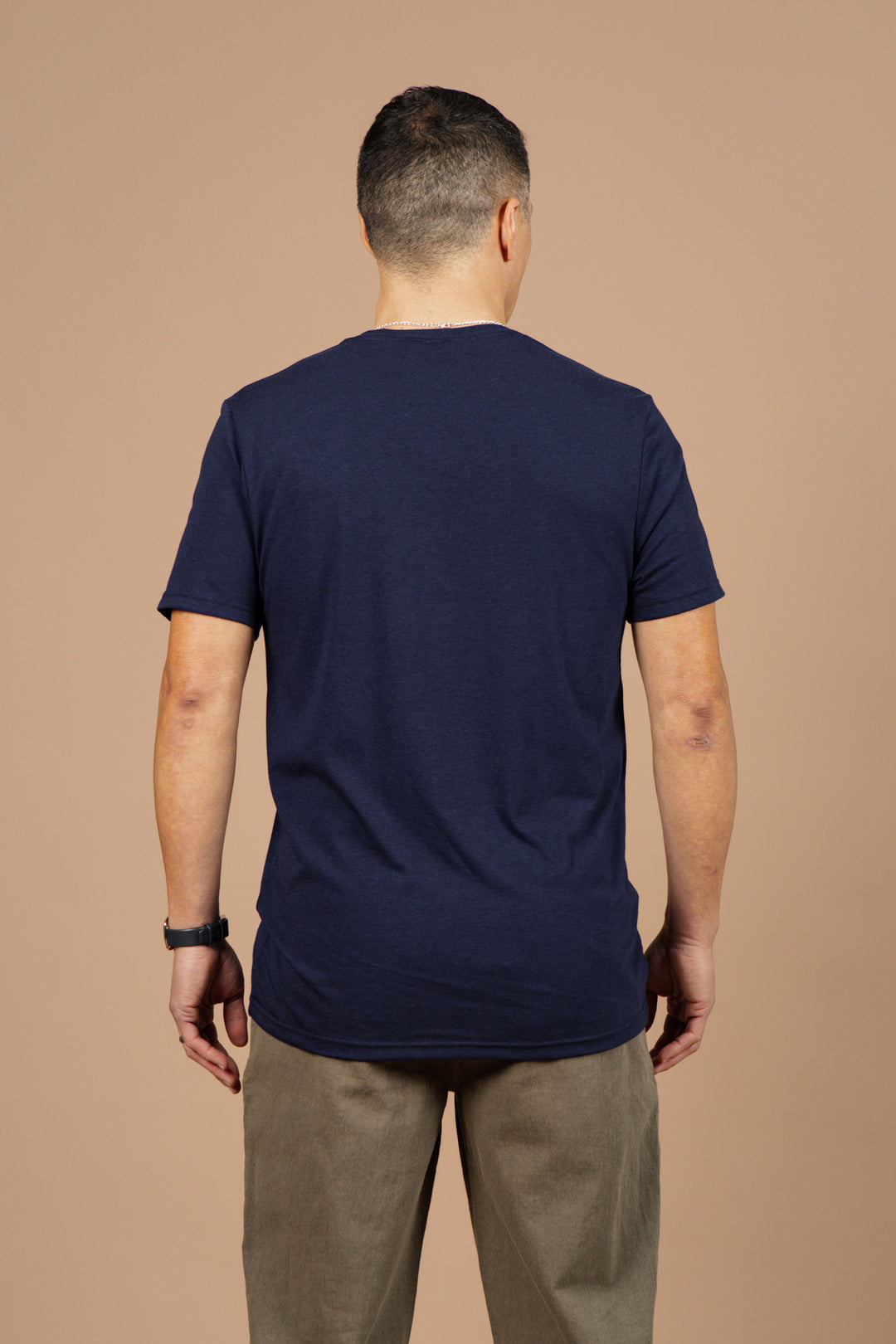 A man stands facing away from the camera, wearing the Bridge & Burn Men's Waves Tee in Navy and khaki pants against a plain beige background.