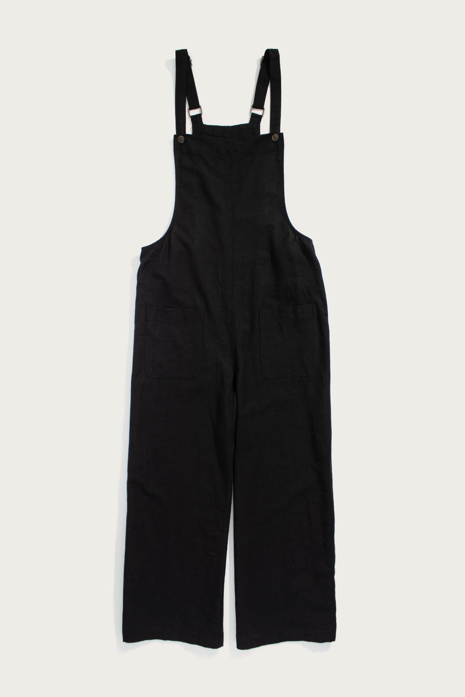 The Edie Overall in Black by Bridge & Burn is a wide-leg style with front pockets and adjustable straps, crafted from a soft tencel-cotton blend and shown laid flat on a white background.