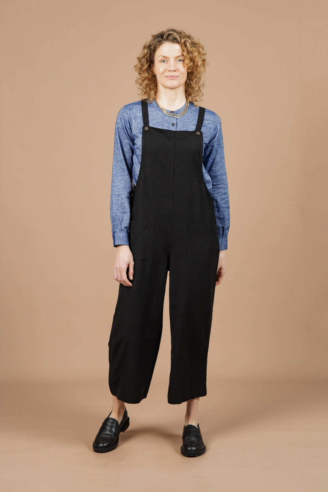 A person with curly hair wears the Bridge & Burn Edie Overall in Black paired with a blue long-sleeve shirt and black shoes, standing and smiling against a plain beige background.