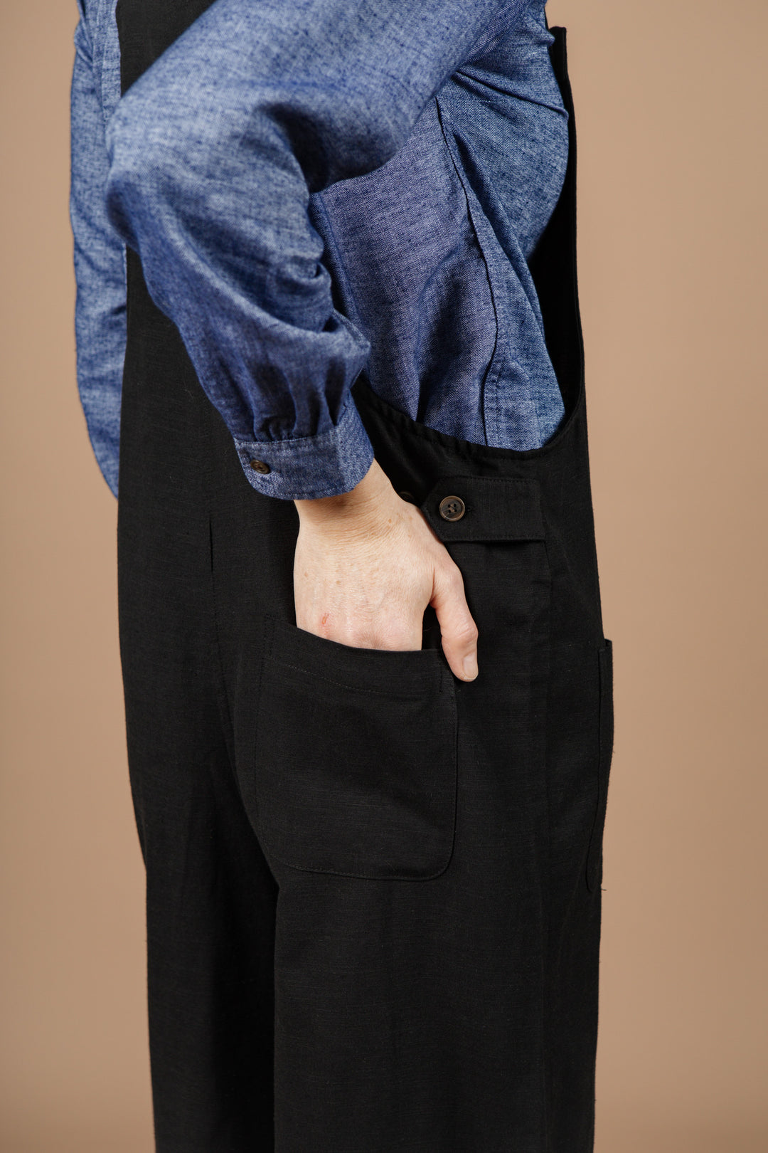 A person wears the Bridge & Burn Edie Overall in black—a wide-leg overall with adjustable straps—paired with a blue long-sleeve shirt. They stand sideways against a plain beige background, their right hand tucked into the side pocket of the overalls.