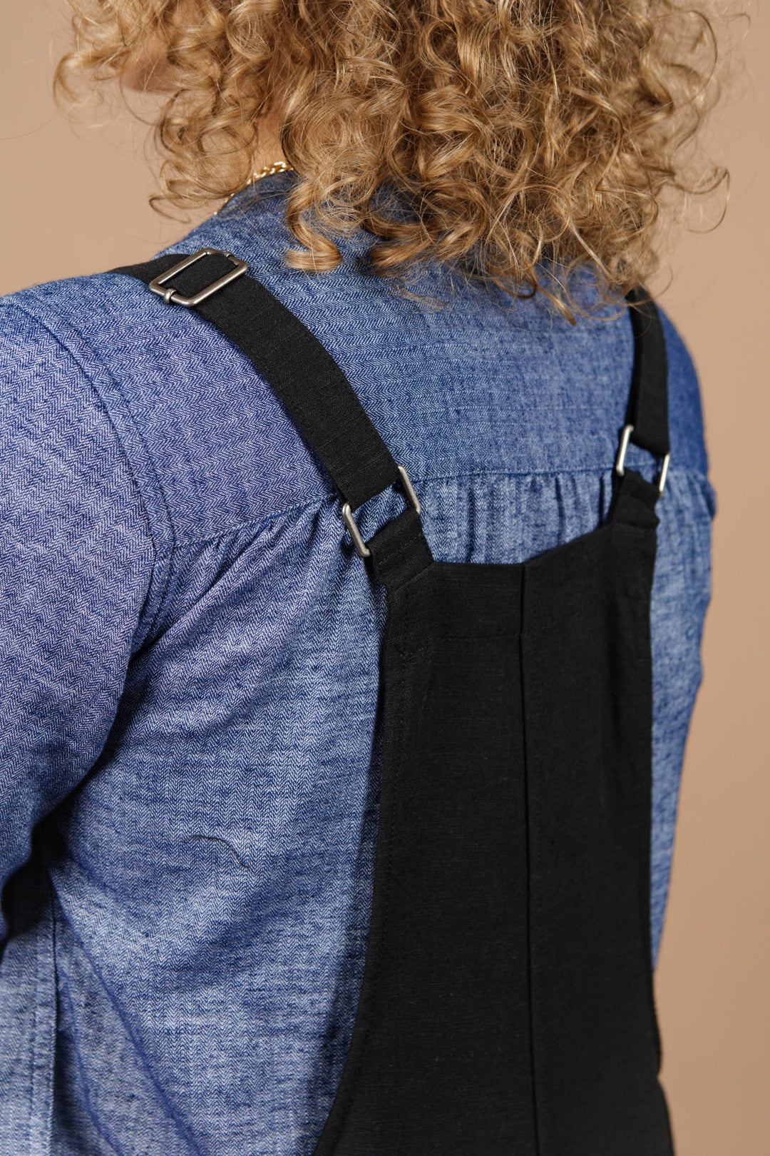 Close-up of a person with curly blonde hair wearing a blue chambray shirt and the Bridge & Burn Edie Overall in black, featuring adjustable straps, viewed from behind.