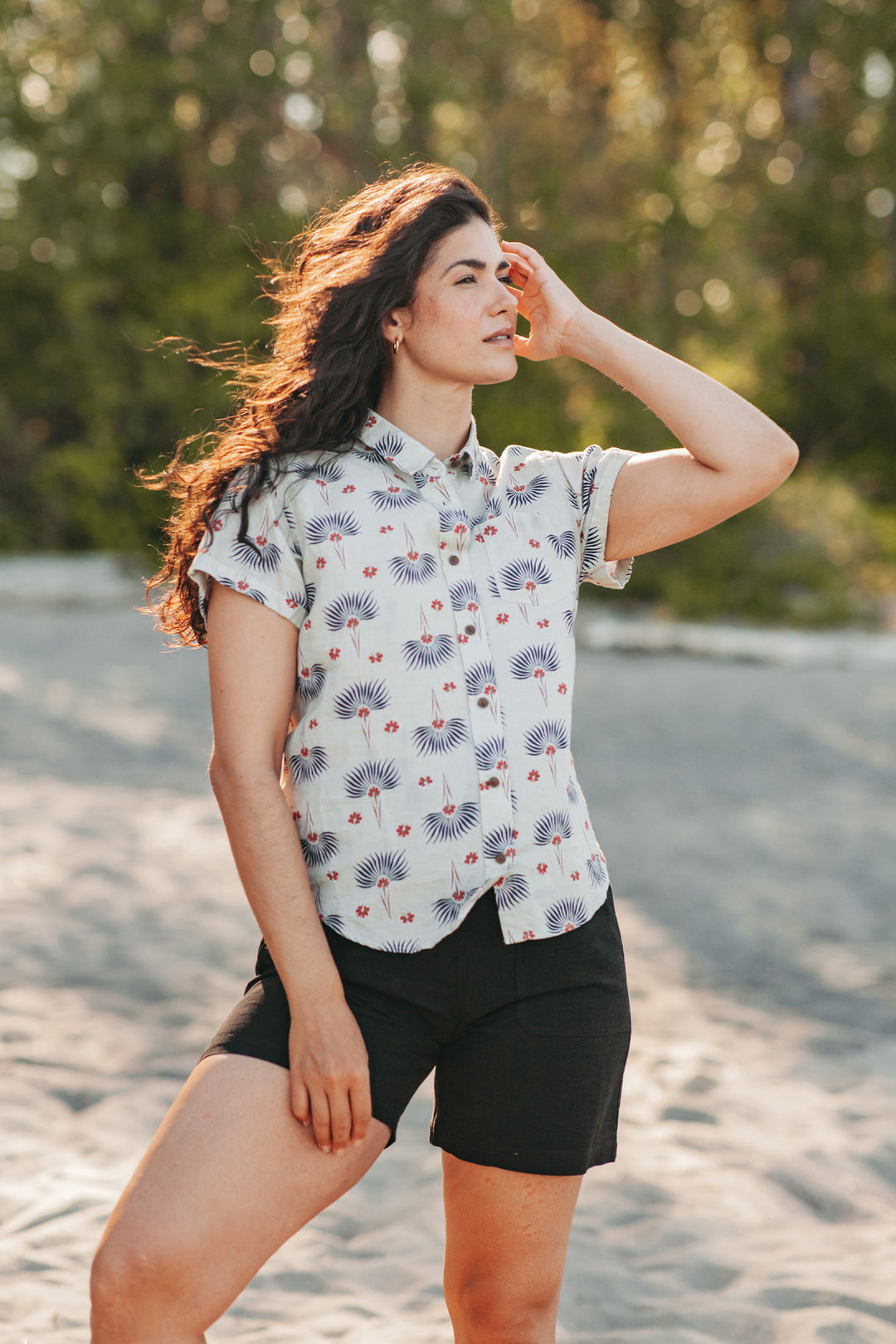 A woman with long wavy hair stands outdoors on sand, sunlight shining on her. She wears the Bridge & Burn Bea Shirt in Aloha Floral and black shorts, with one hand touching her hair. Trees form a natural backdrop.