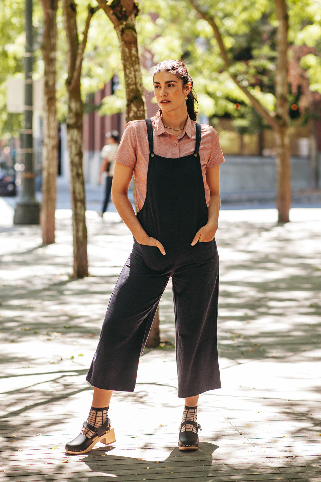 A woman stands outdoors on a sunlit sidewalk, wearing the Bridge & Burn Edie Overall in Navy over a pink shirt and black sandals. Trees and buildings appear in the background.