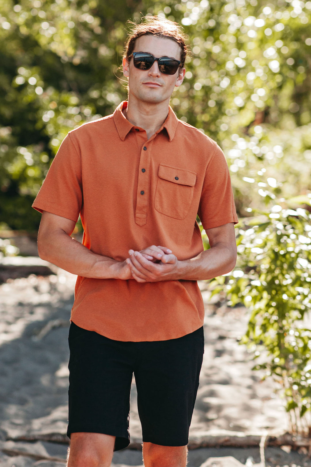 A man stands outdoors on a sunny day wearing Bridge & Burn’s Gary Knit Pullover in Clay Red Jersey and black shorts, with green trees and sunlight softly blurred in the background.
