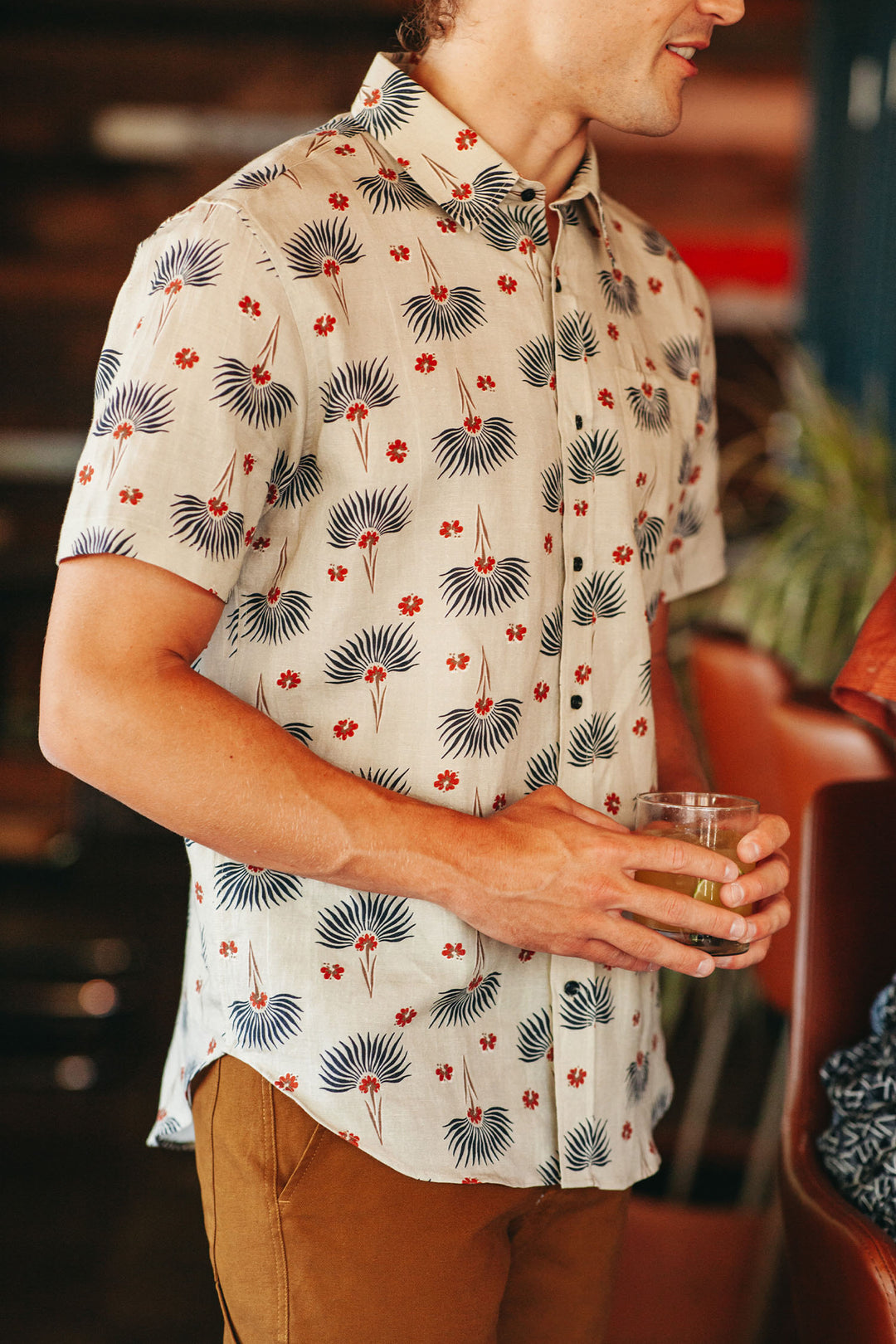 Wearing the Bridge & Burn Harbor Slim Shirt in Aloha Floral, a person stands indoors by brown furniture, holding a glass. The white, short-sleeve, slim-fit shirt with blue and red floral pattern is partially visible; their face is partly out of frame.